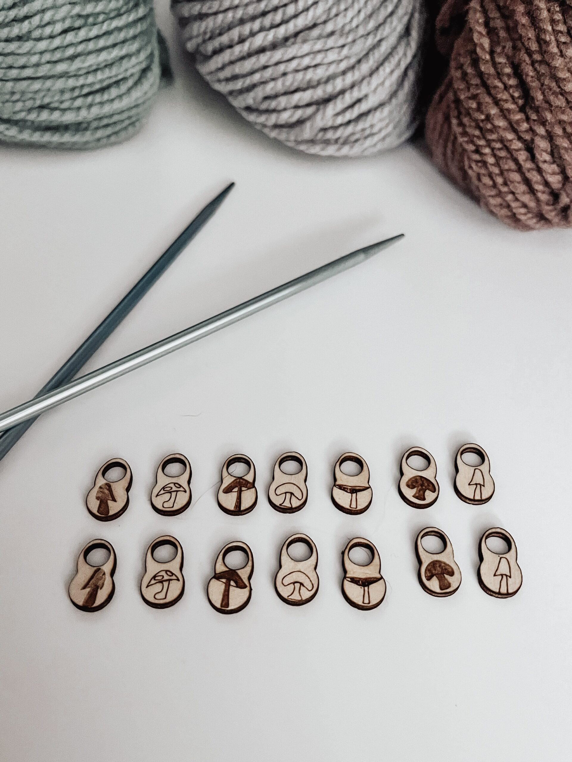 A total of 14 wood stitch markers with a variety of hand-drawn mushroom designs are displayed with a pair of metal knitting needles and three skeins of merino yarn are in the background.