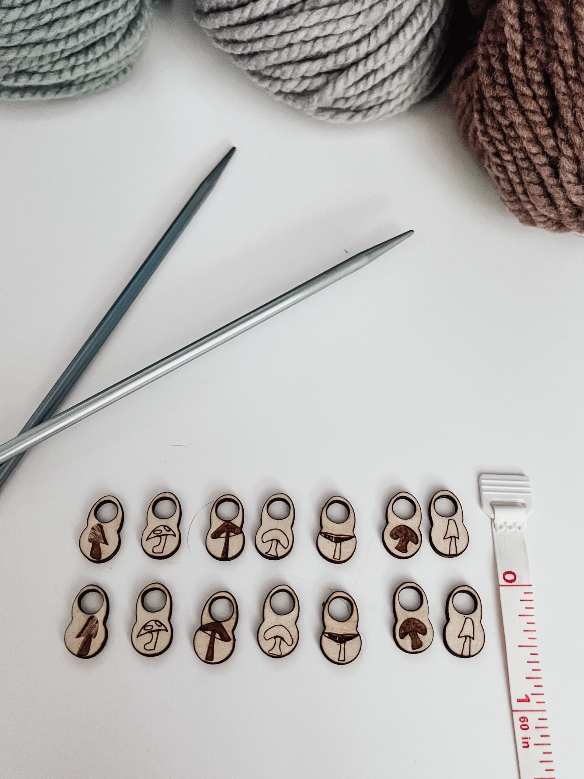 A total of 14 wood stitch markers with a variety of hand-drawn mushroom designs are displayed next to a measuring tape that shows they're approximately 1/2" tall. A pair of metal knitting needles and three skeins of merino yarn are in the background.
