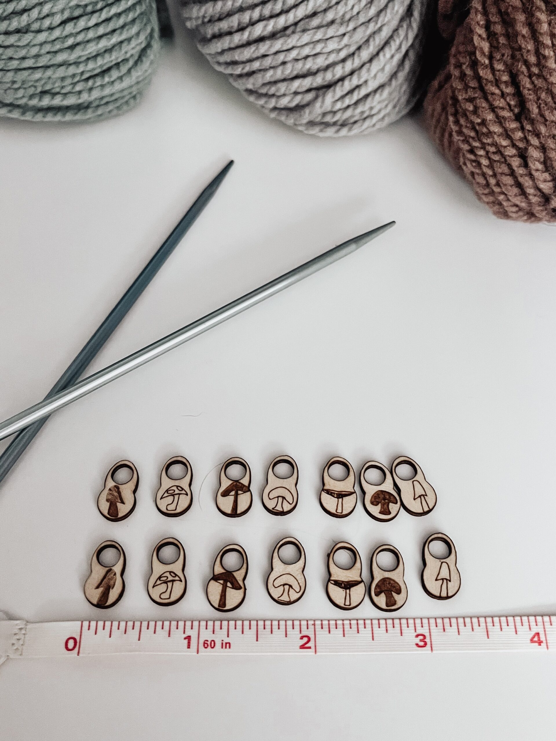 A total of 14 wood stitch markers with a variety of hand-drawn mushroom designs are displayed next to a measuring tape that shows they're approximately 1/4" wide. A pair of metal knitting needles and three skeins of merino yarn are in the background.