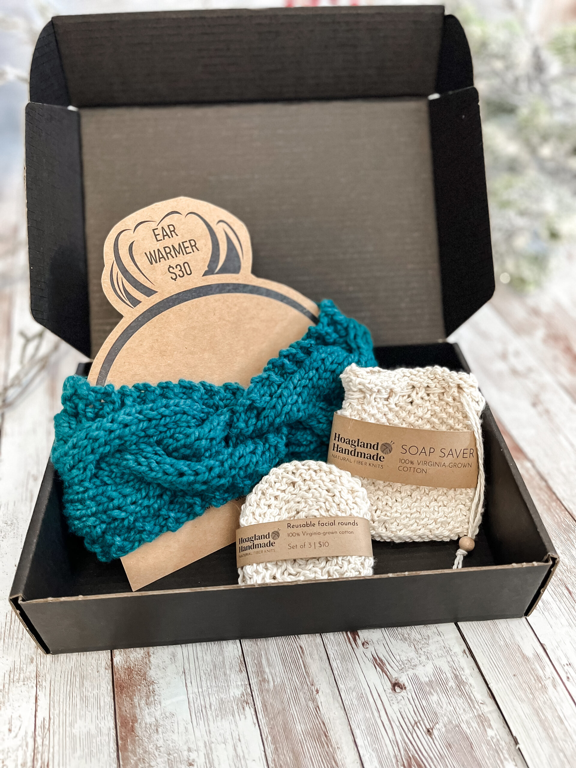 A black box contains a teal hand-dyed merino ear warmer displayed on a Kraft paper head with a messy bun, a Virginia-grown cotton soap saver and set of reusable facial rounds.