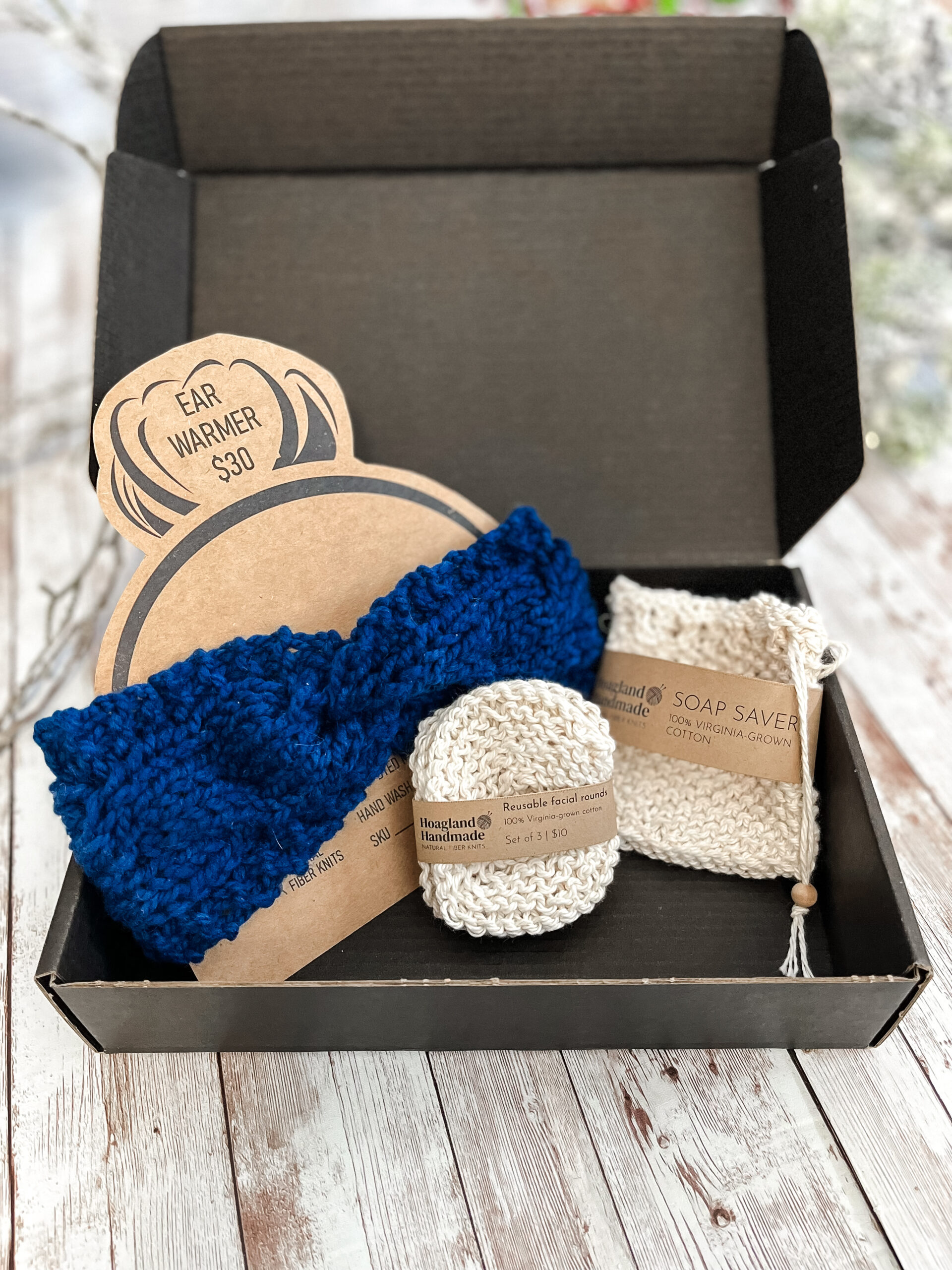 A black box contains a blue hand-dyed merino ear warmer displayed on a Kraft paper head with a messy bun, a Virginia-grown cotton soap saver and set of reusable facial rounds.