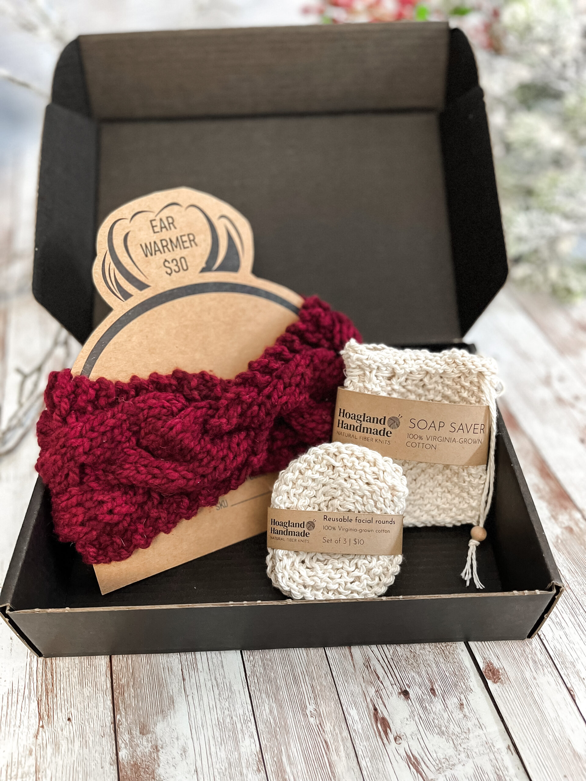 A black box contains a red hand-dyed merino ear warmer displayed on a Kraft paper head with a messy bun, a Virginia-grown cotton soap saver and set of reusable facial rounds.