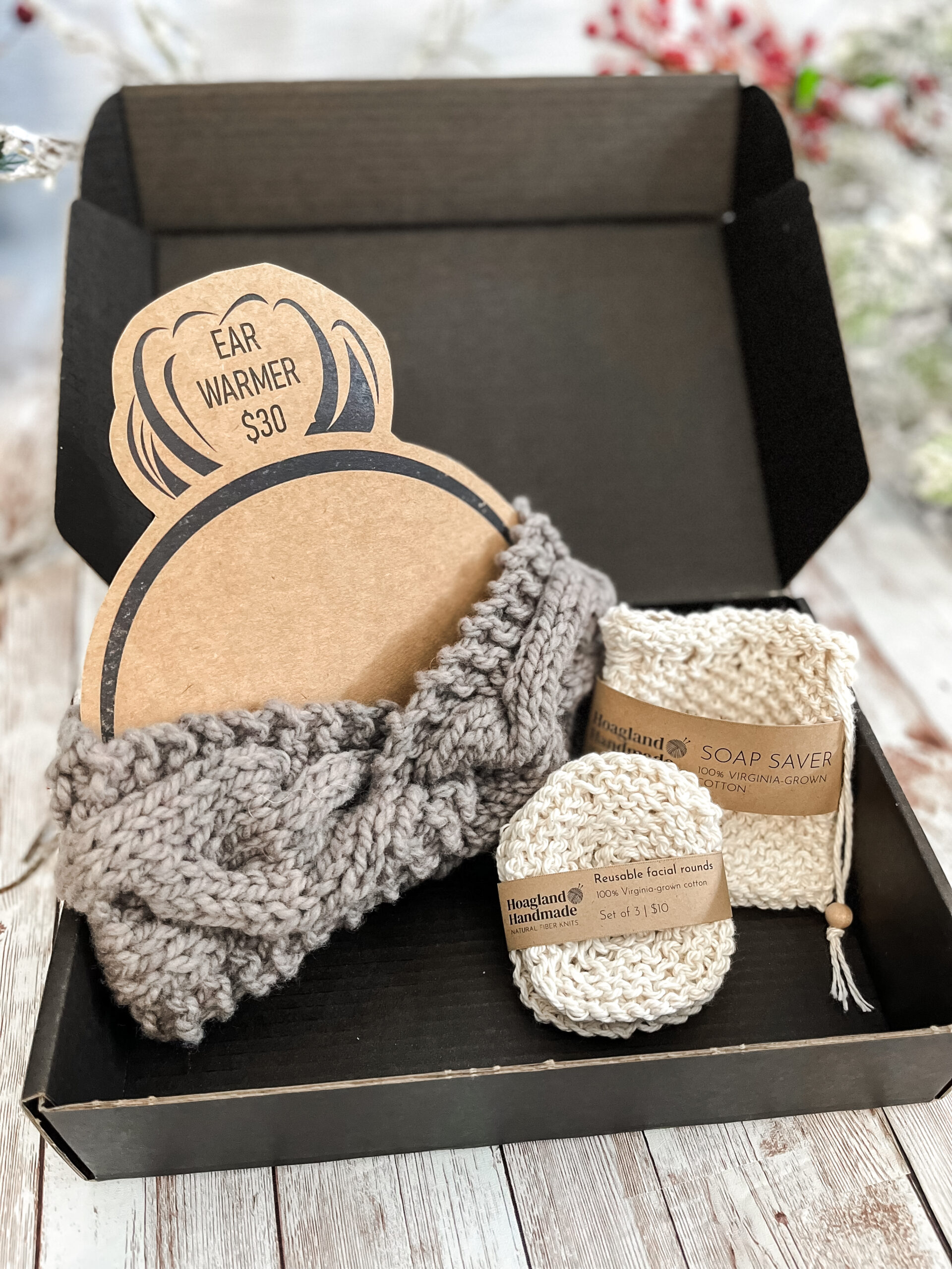 A black box contains a gray hand-dyed merino ear warmer displayed on a Kraft paper head with a messy bun, a Virginia-grown cotton soap saver and set of reusable facial rounds.