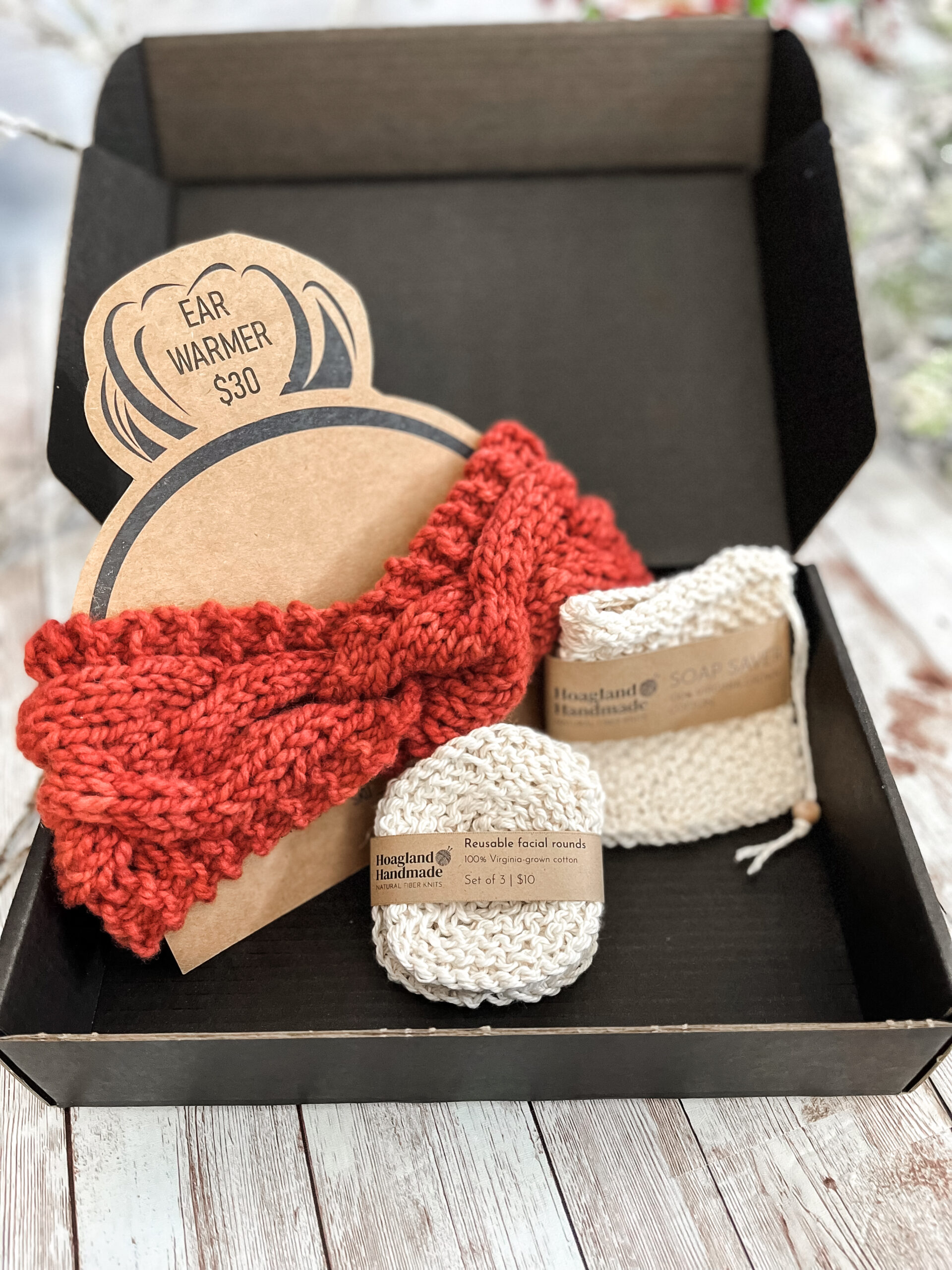 A black box contains an orange hand-dyed merino ear warmer displayed on a Kraft paper head with a messy bun, a Virginia-grown cotton soap saver and set of reusable facial rounds.