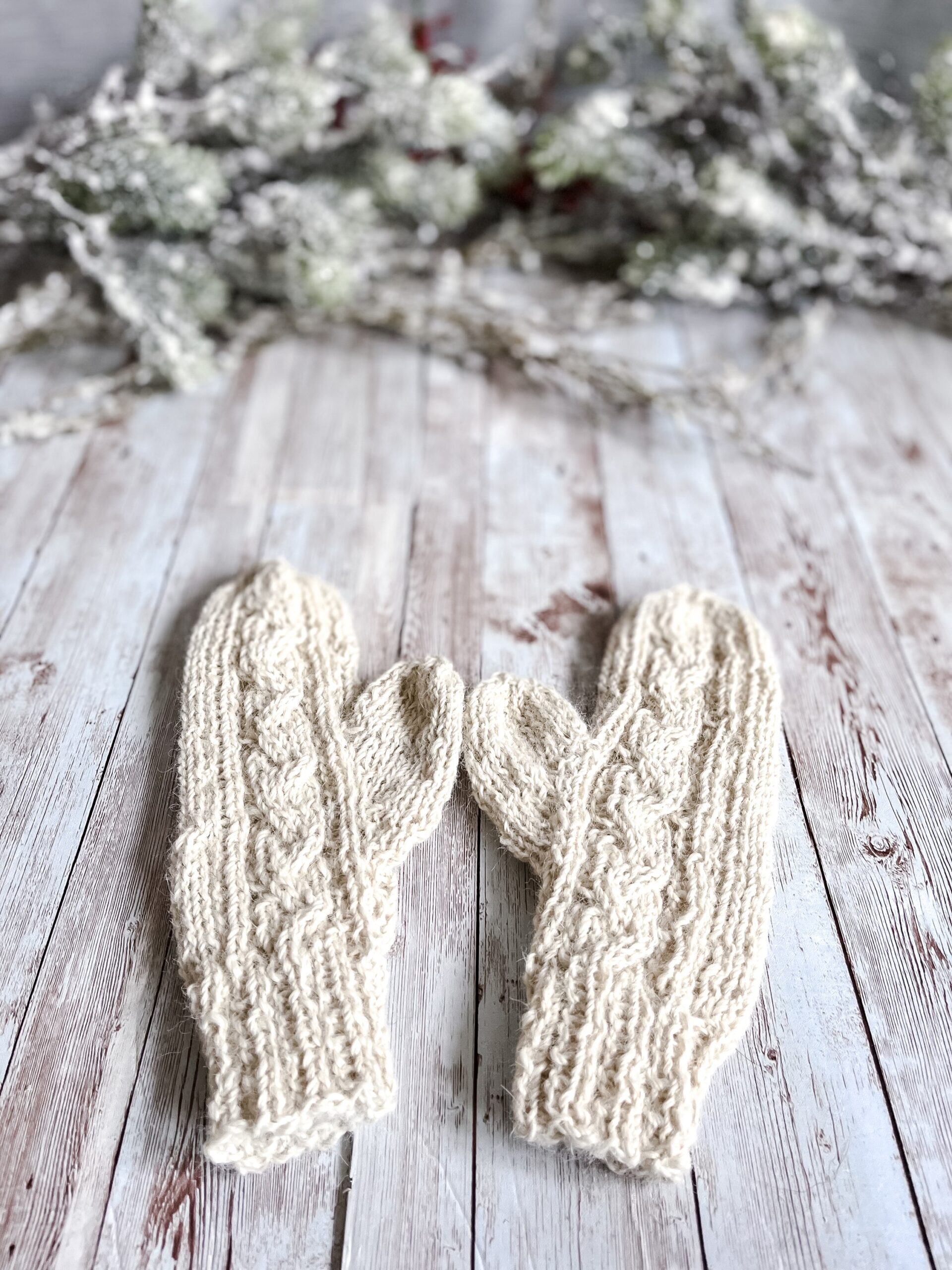 A pair of natural white alpaca mittens rests on a wood plank background. In the background of the photo is snow covered pine branches