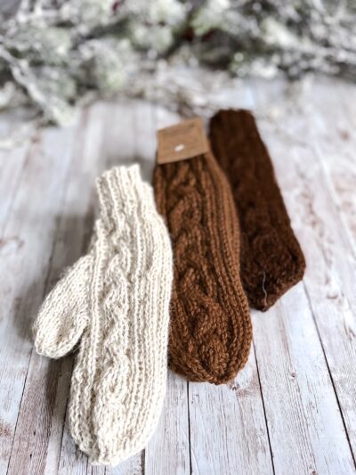 Three pairs of alpaca mittens in natural dark brown, medium brown and white rests on a wood plank background. In the background of the photo is snow covered pine branches