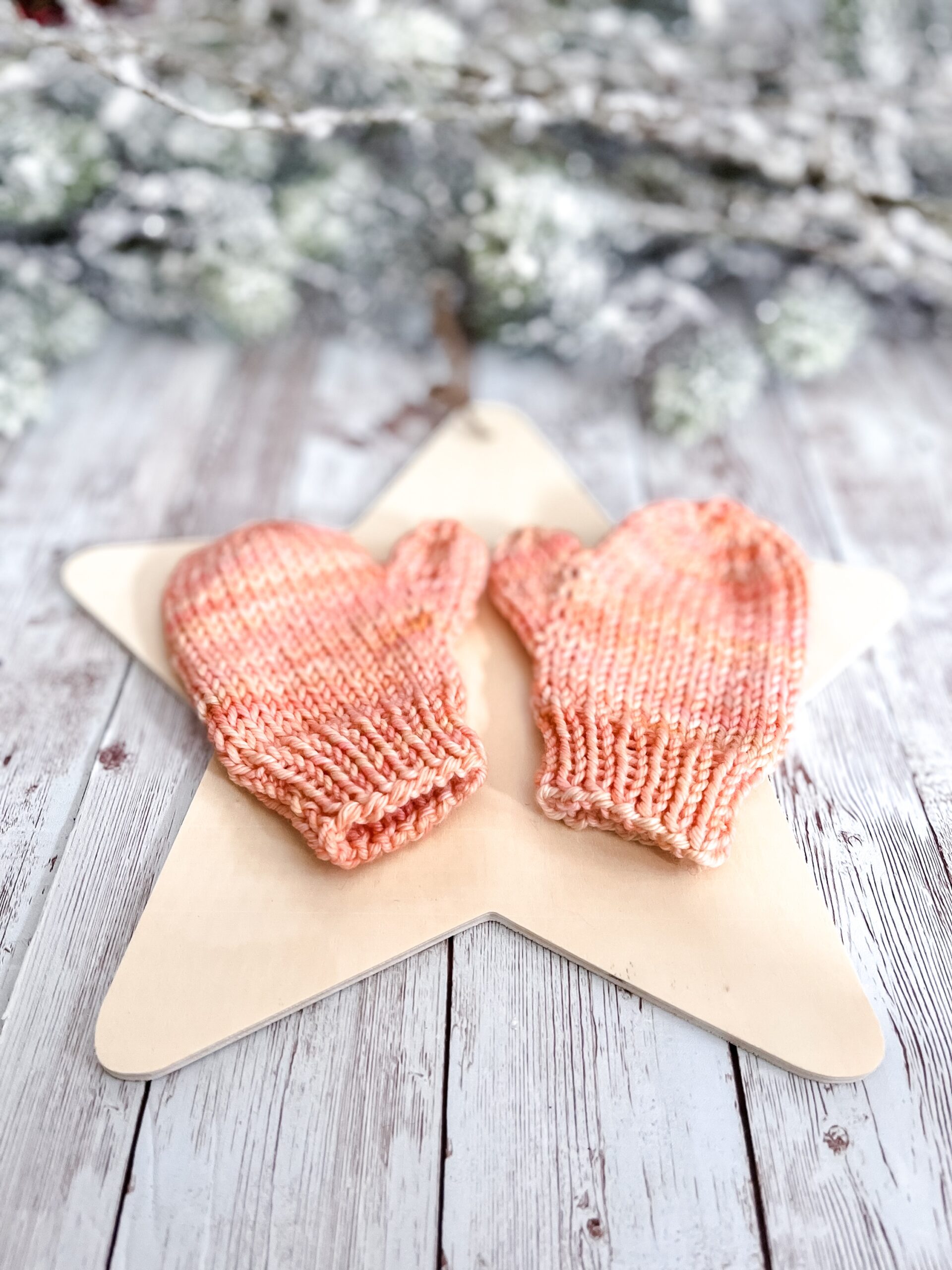 A pair of peach colored mittens rests on a wooden star cutout on a wood plank background. In the background of the photo is snow covered pine branches