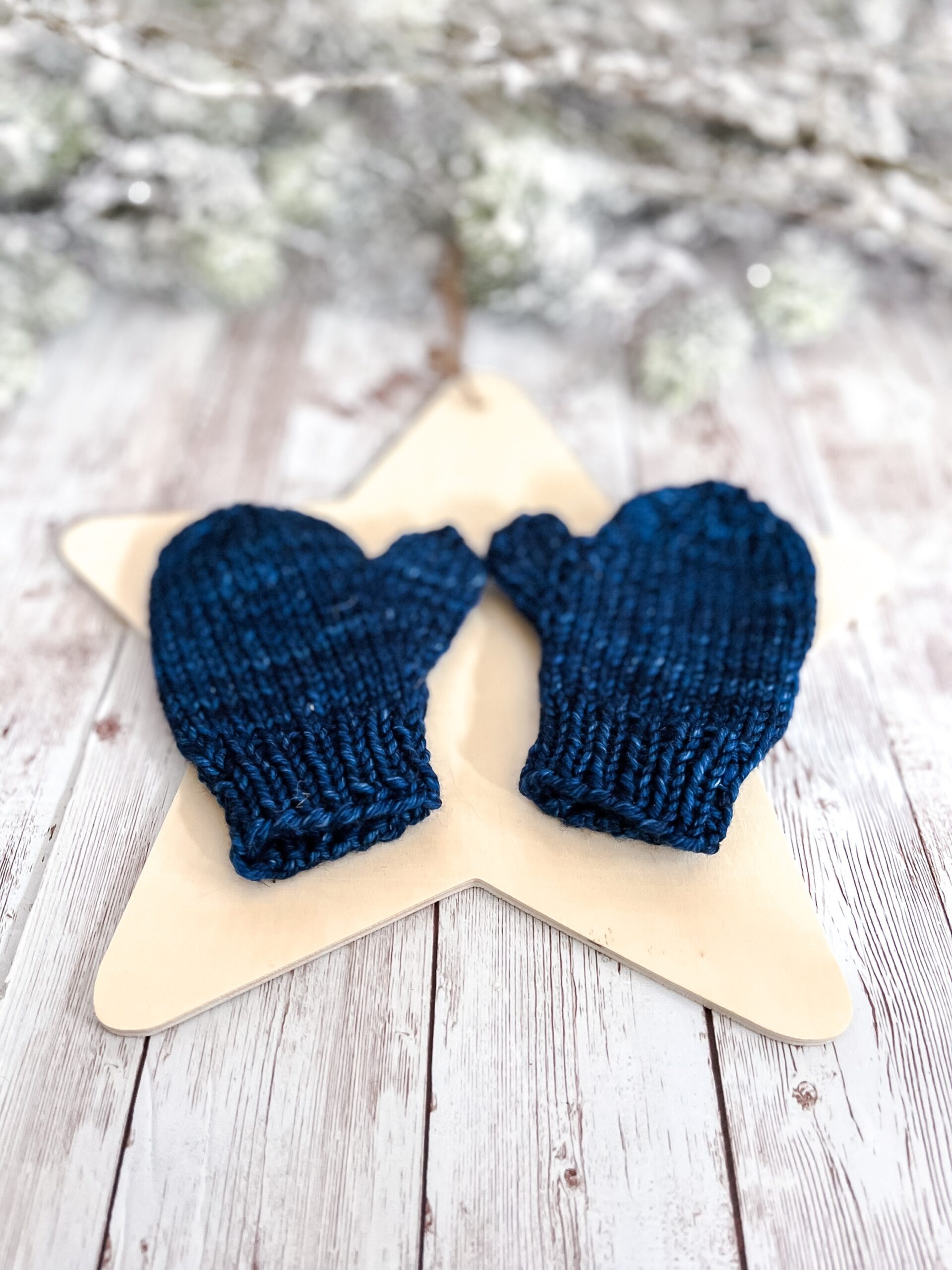 A pair of dark blue colored mittens rests on a wooden star cutout on a wood plank background. In the background of the photo is snow covered pine branches