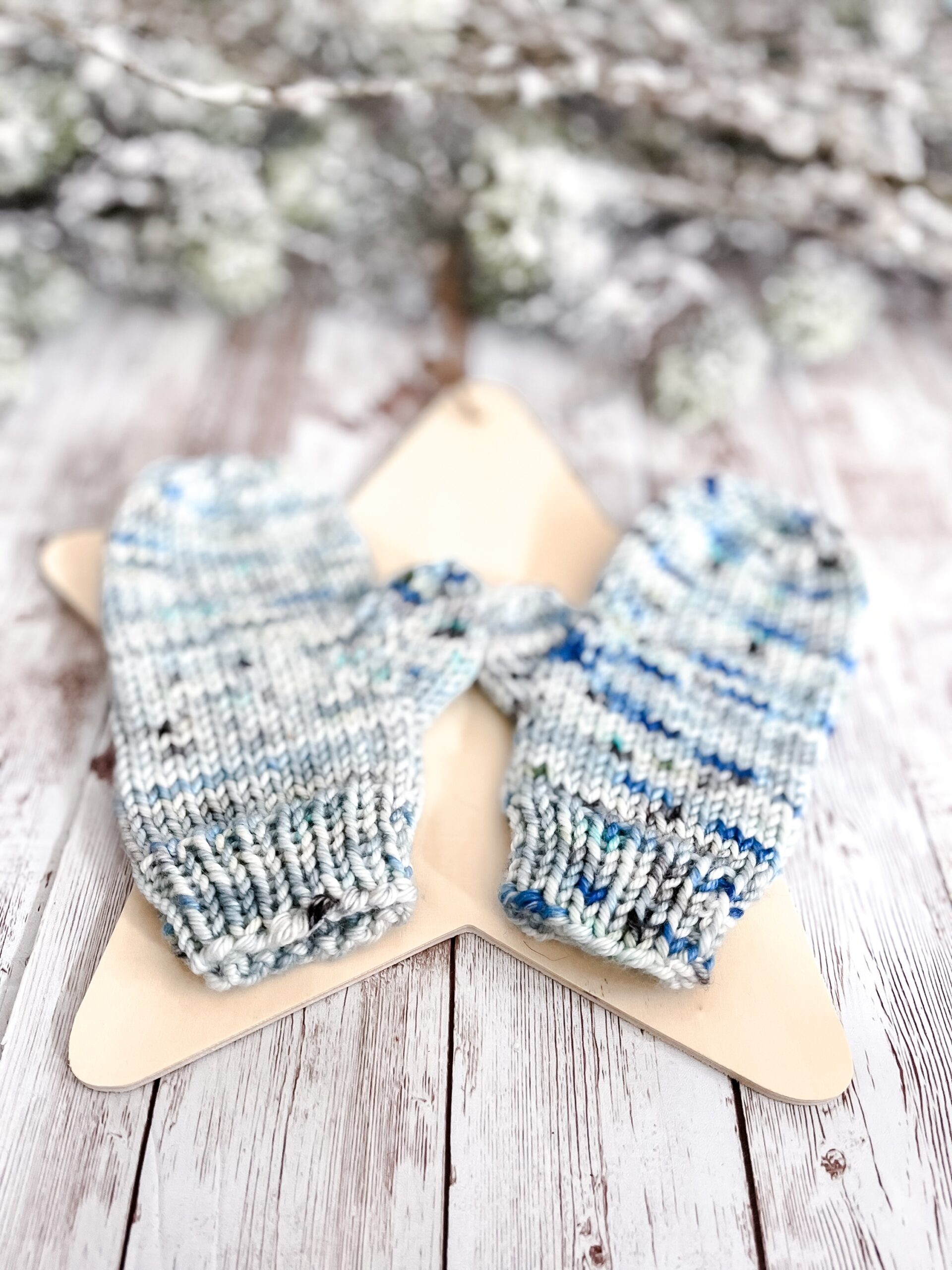 A pair of blue speckled colored mittens rests on a wooden star cutout on a wood plank background. In the background of the photo is snow covered pine branches