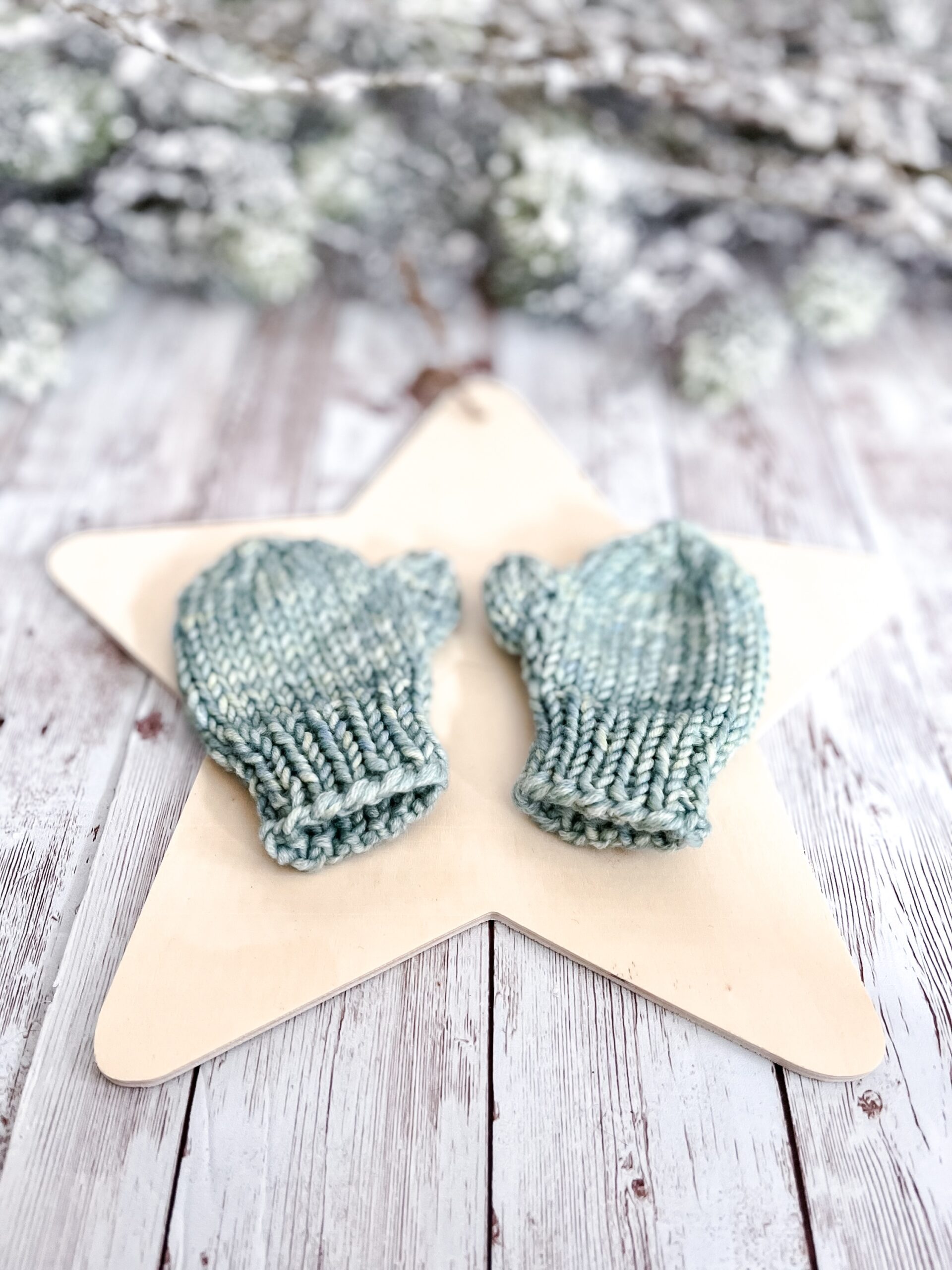 A pair of light green colored mittens rests on a wooden star cutout on a wood plank background. In the background of the photo is snow covered pine branches