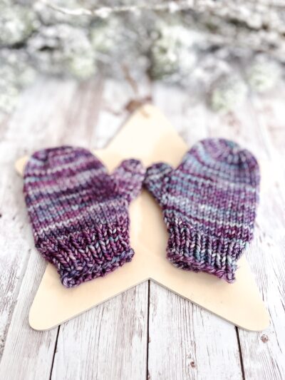 A pair of purple & teal colored mittens rests on a wooden star cutout on a wood plank background. In the background of the photo is snow covered pine branches