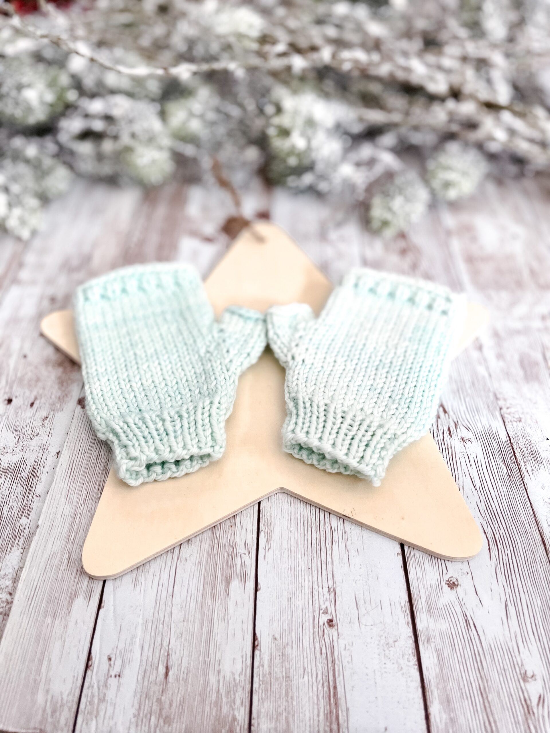 A pair of dark blue fingerless mittens rests on a wooden star cutout on a wood plank background. In the background of the photo is snow covered pine branches