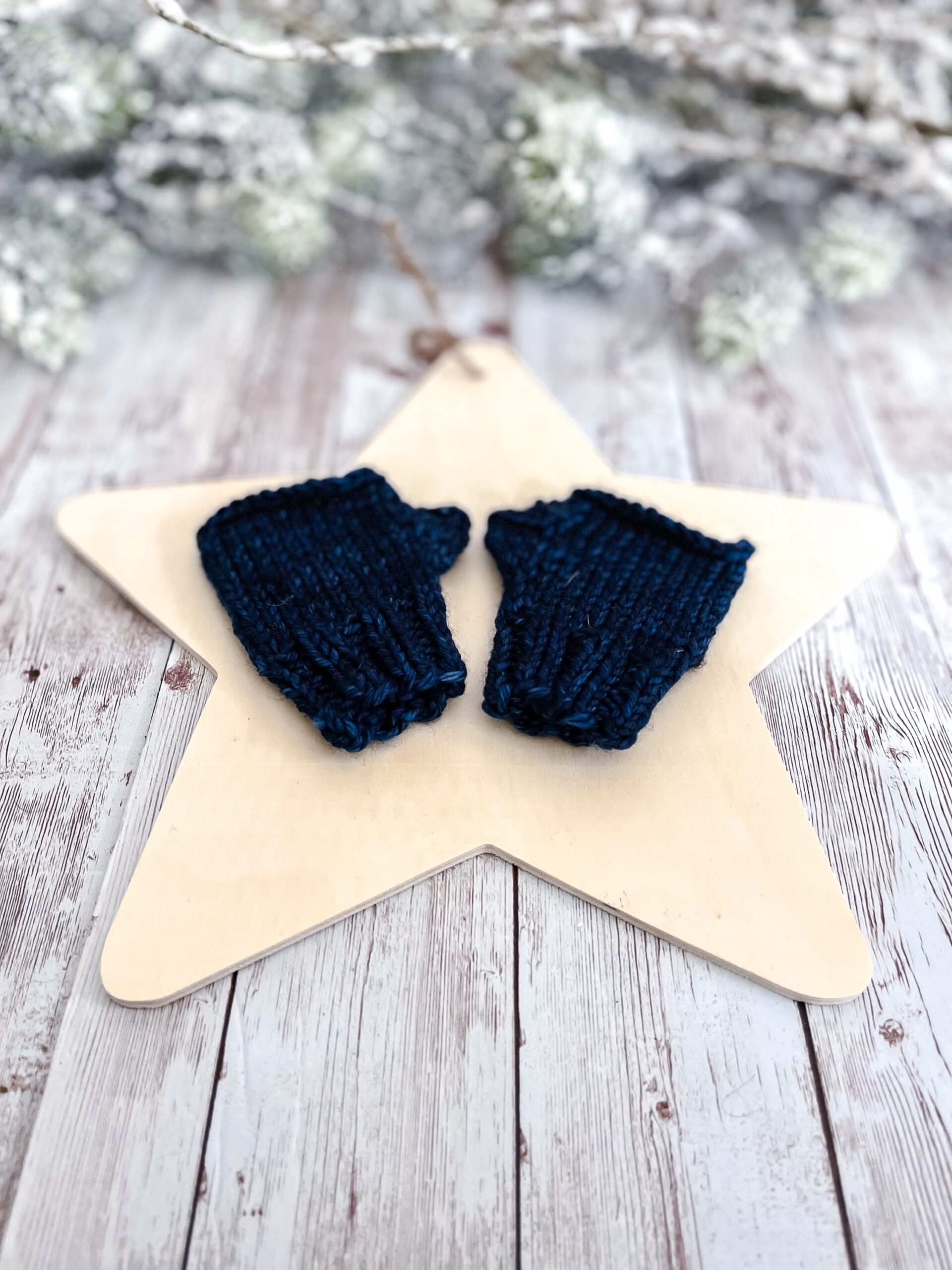 A pair of dark blue fingerless mittens rests on a wooden star cutout on a wood plank background. In the background of the photo is snow covered pine branches