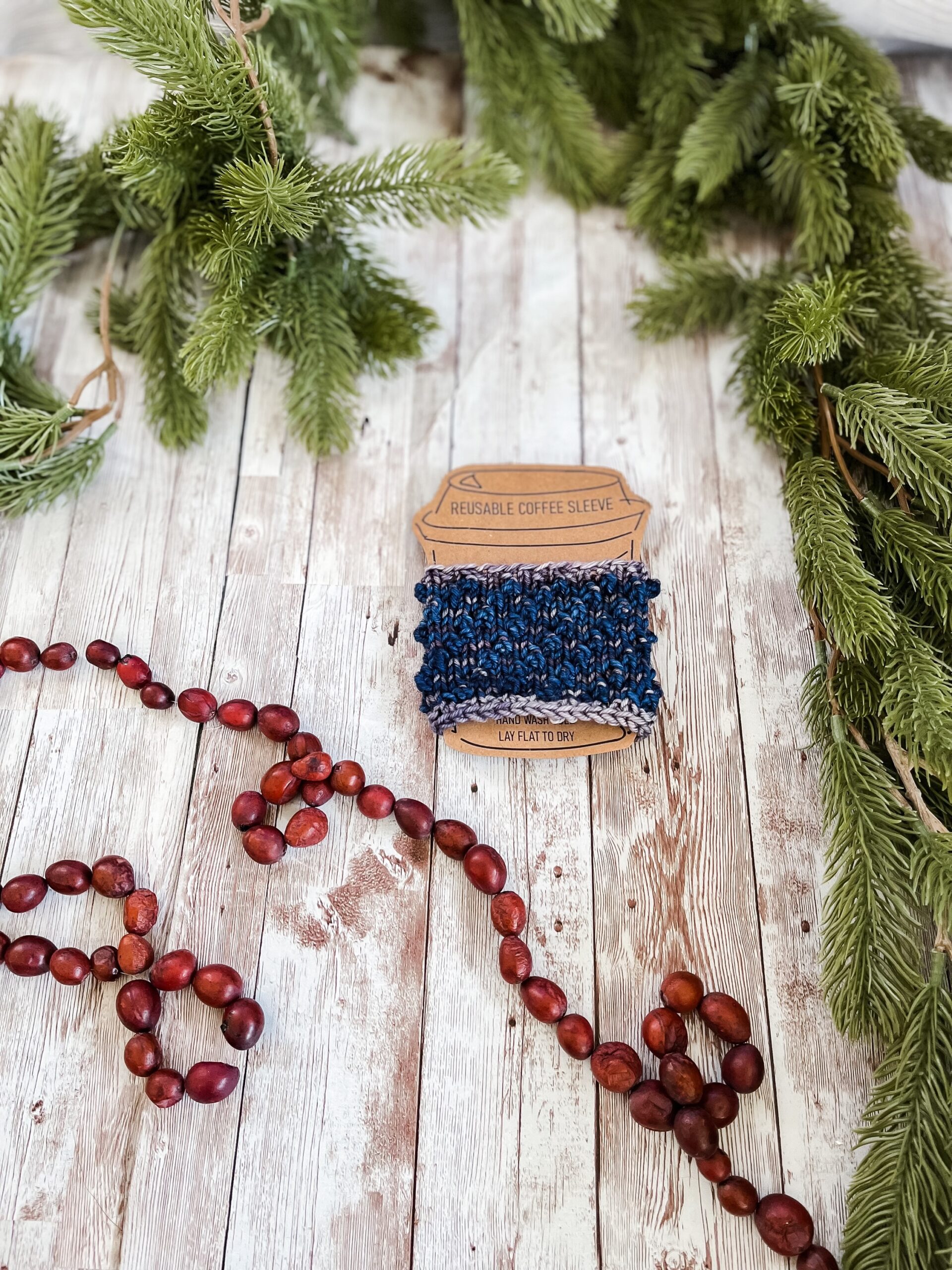 A blue and gray coffee sleeve is wrapped around a kraft paper image of a coffee cup. It rests on a wood plank, surrounded by pine branches and a cranberry garland.