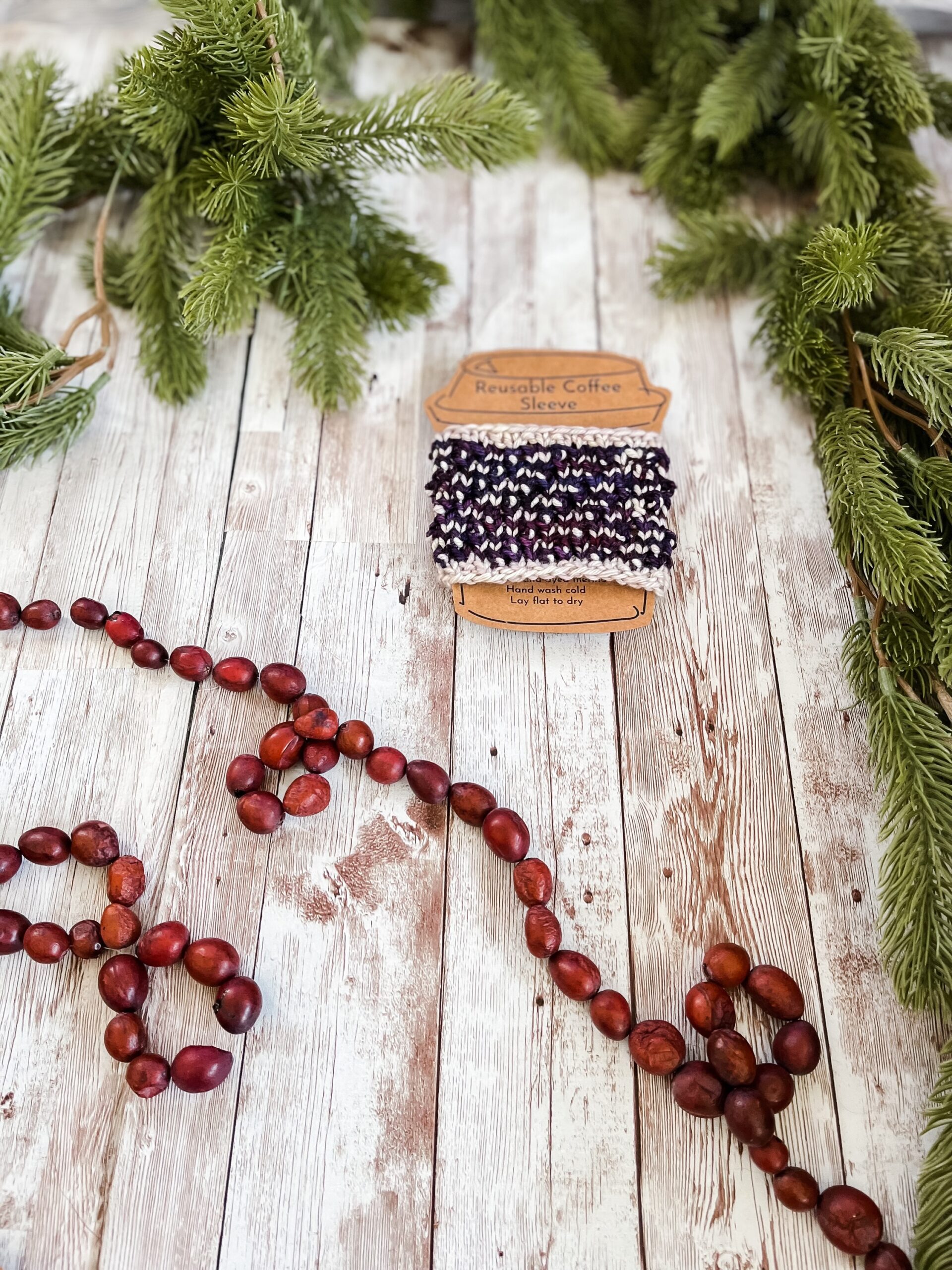 A purple and white coffee sleeve is wrapped around a kraft paper image of a coffee cup. It rests on a wood plank, surrounded by pine branches and a cranberry garland.