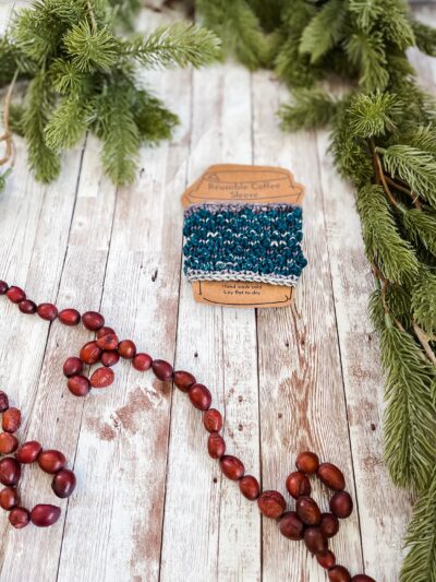 A teal and gray coffee sleeve is wrapped around a kraft paper image of a coffee cup. It rests on a wood plank, surrounded by pine branches and a cranberry garland.