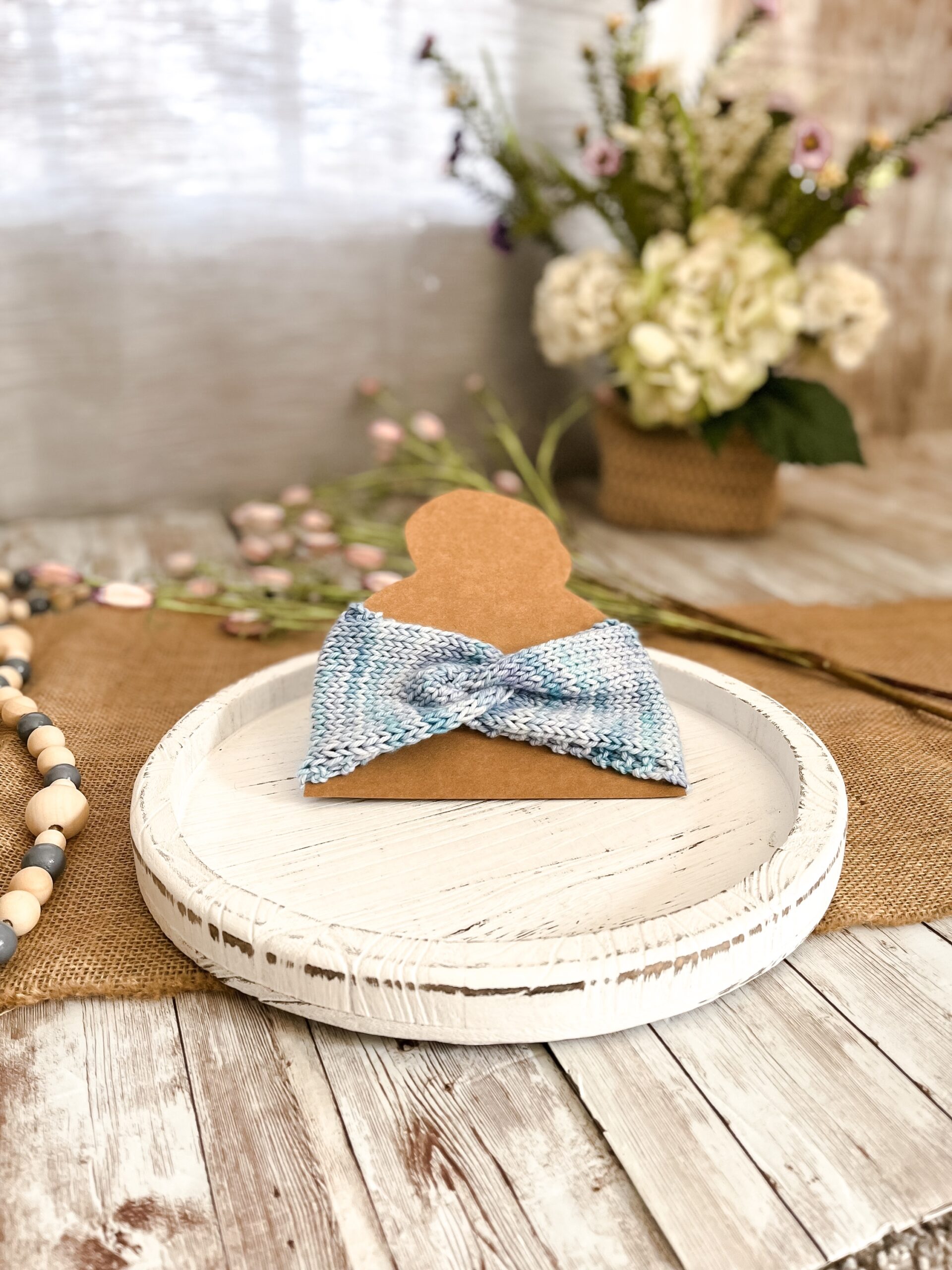 a light blue twisted ear warmer is displayed on a kraft paper cut out of a head with a top knot. In the background is a spring floral arrangement, some beads and some flowers laying on a burlap table runner resting on a wood plank