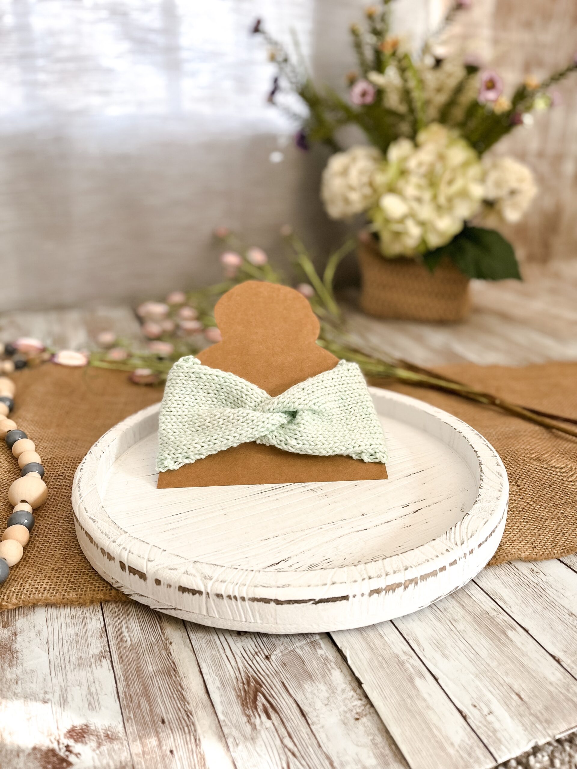 a pale green twisted ear warmer is displayed on a kraft paper cut out of a head with a top knot. In the background is a spring floral arrangement, some beads and some flowers laying on a burlap table runner resting on a wood plank