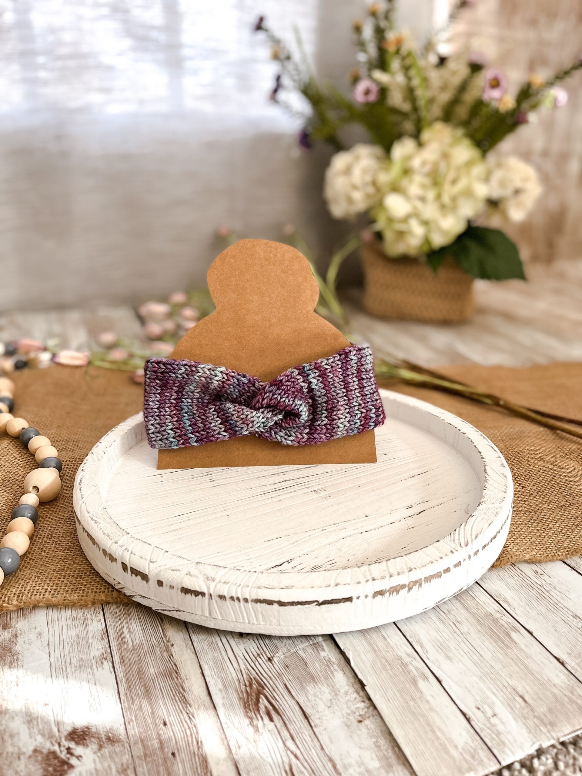 a teal and purple twisted ear warmer is displayed on a kraft paper cut out of a head with a top knot. In the background is a spring floral arrangement, some beads and some flowers laying on a burlap table runner resting on a wood plank
