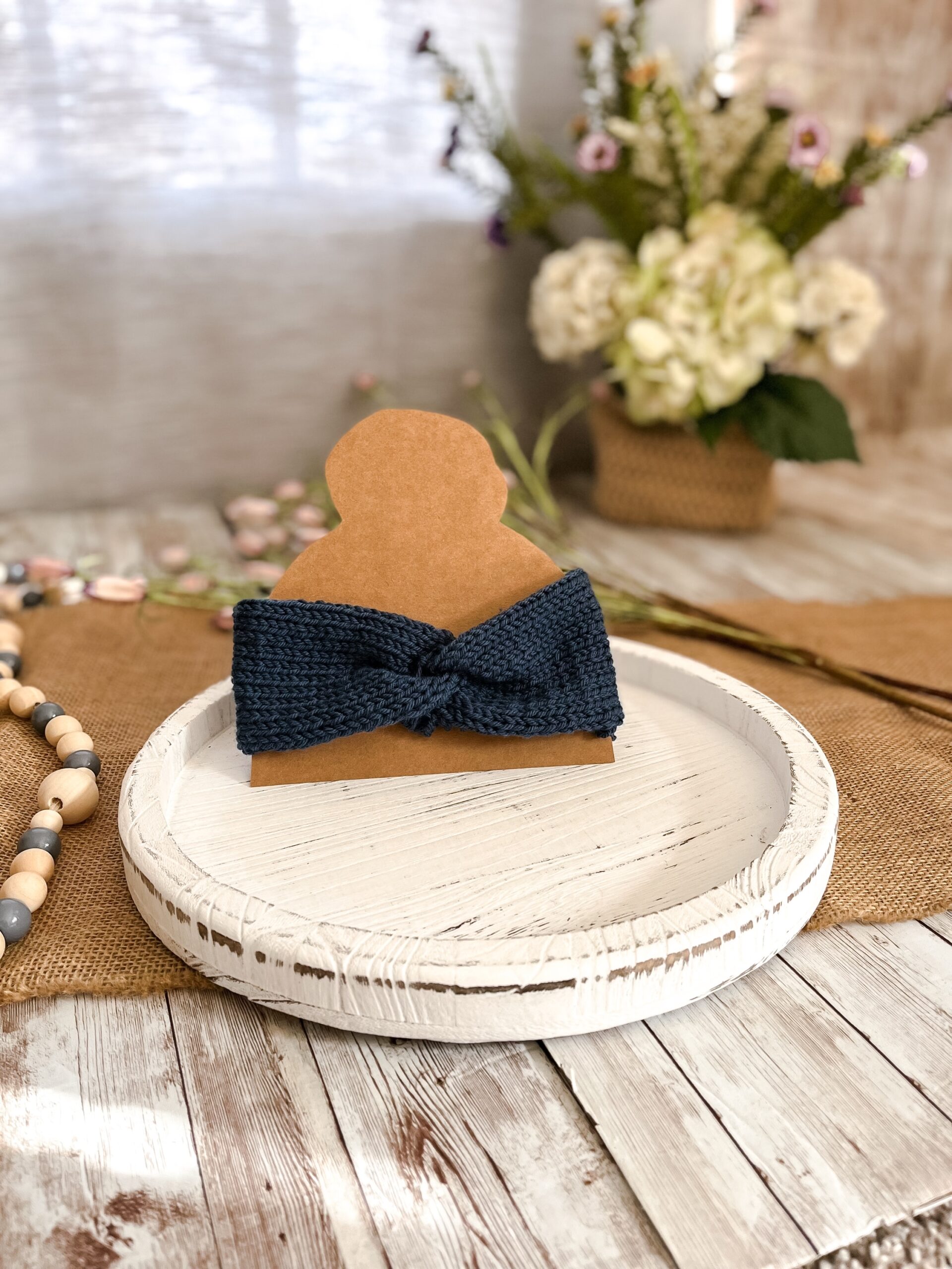 a denim blue twisted ear warmer is displayed on a kraft paper cut out of a head with a top knot. In the background is a spring floral arrangement, some beads and some flowers laying on a burlap table runner resting on a wood plank