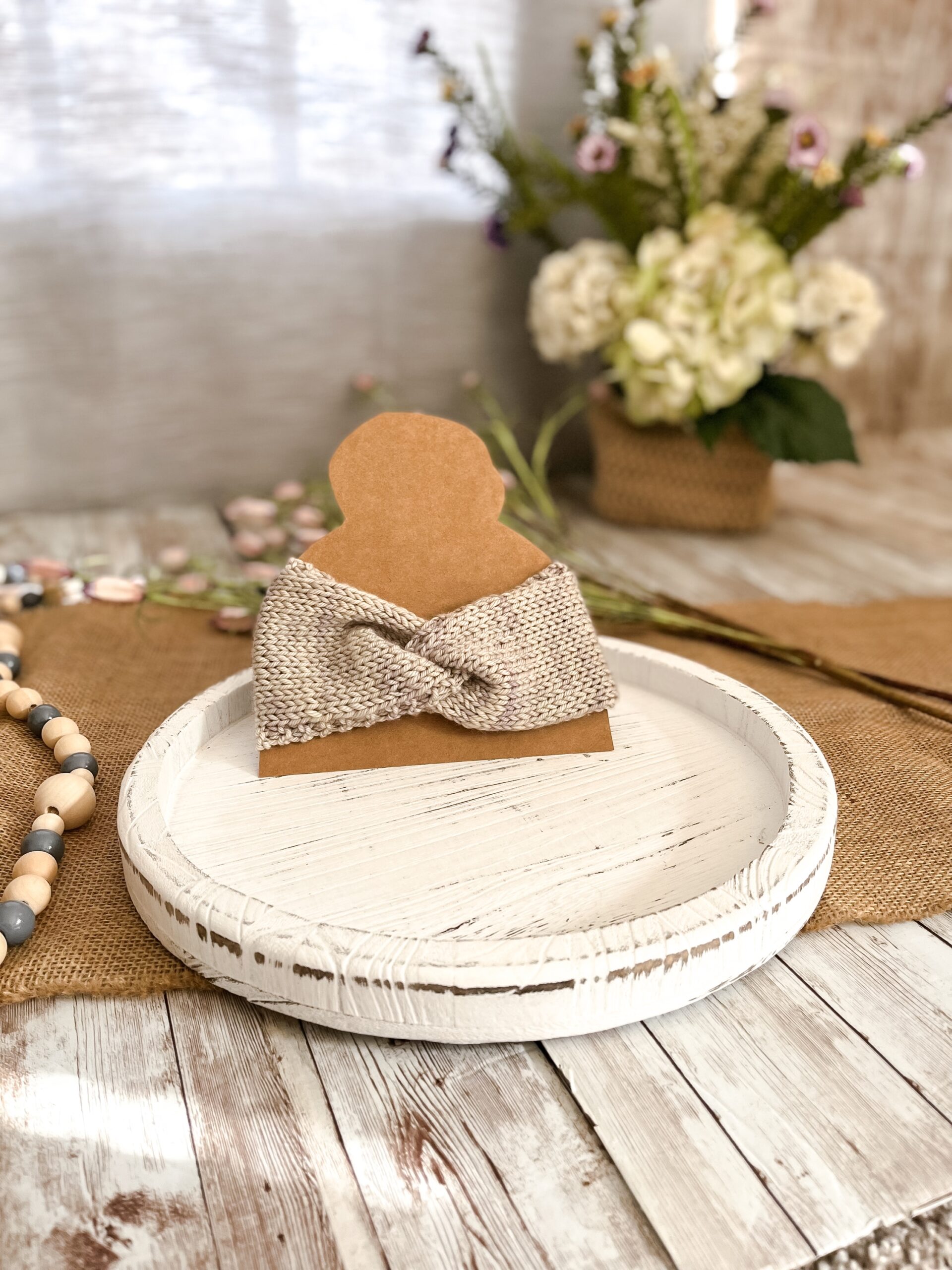 a light gray twisted ear warmer is displayed on a kraft paper cut out of a head with a top knot. In the background is a spring floral arrangement, some beads and some flowers laying on a burlap table runner resting on a wood plank