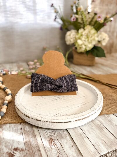a blueish gray twisted ear warmer is displayed on a kraft paper cut out of a head with a top knot. In the background is a spring floral arrangement, some beads and some flowers laying on a burlap table runner resting on a wood plank