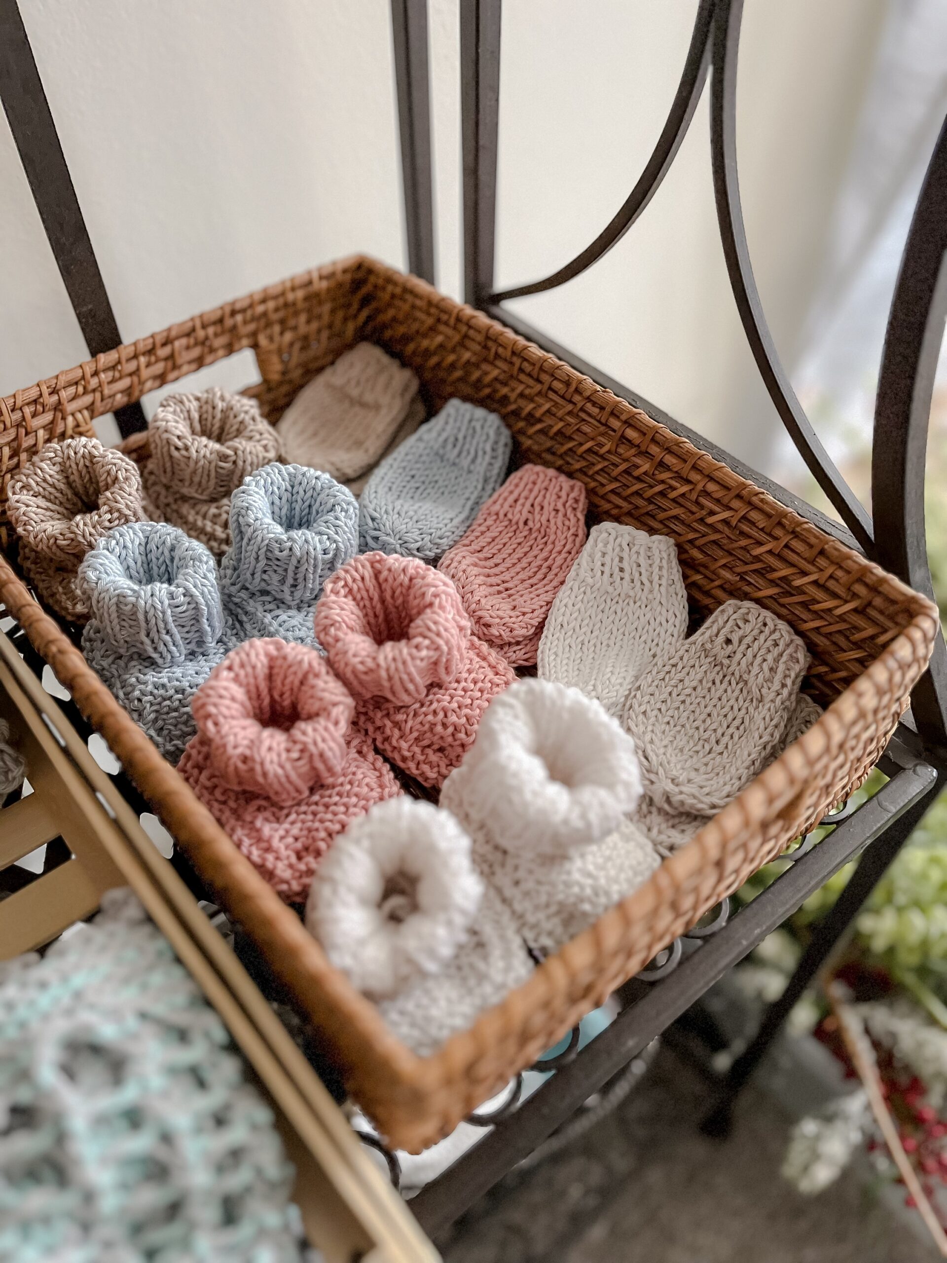 A wicker basket holds organic cotton newborn booties and mitts. The booties are tan, blue, pink, and white, and the mitts are tan, blue, pink, white, and gray.