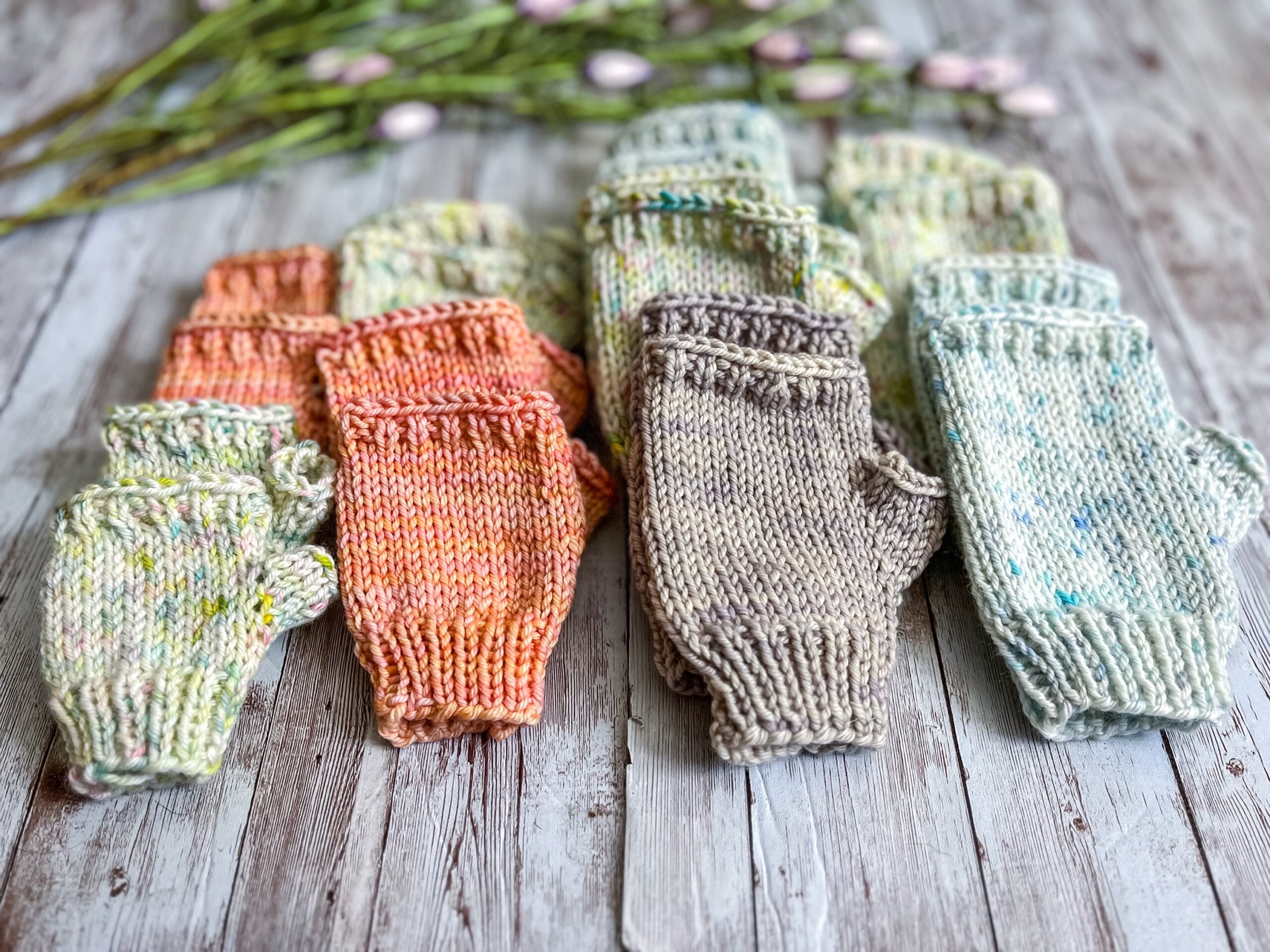 Multiple pairs of hand-dyed merino fingerless mitts in a variety of children's sizes and spring colors are displayed with flowers in the background. Colors of the fingerless mitts include a pale yellow speckle, pale blue speckle, a peach tonal, and a light gray tonal