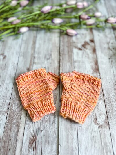 A pair of kid's sized peach colored fingerless mitts are displayed on a wood plank with flower buds in the background