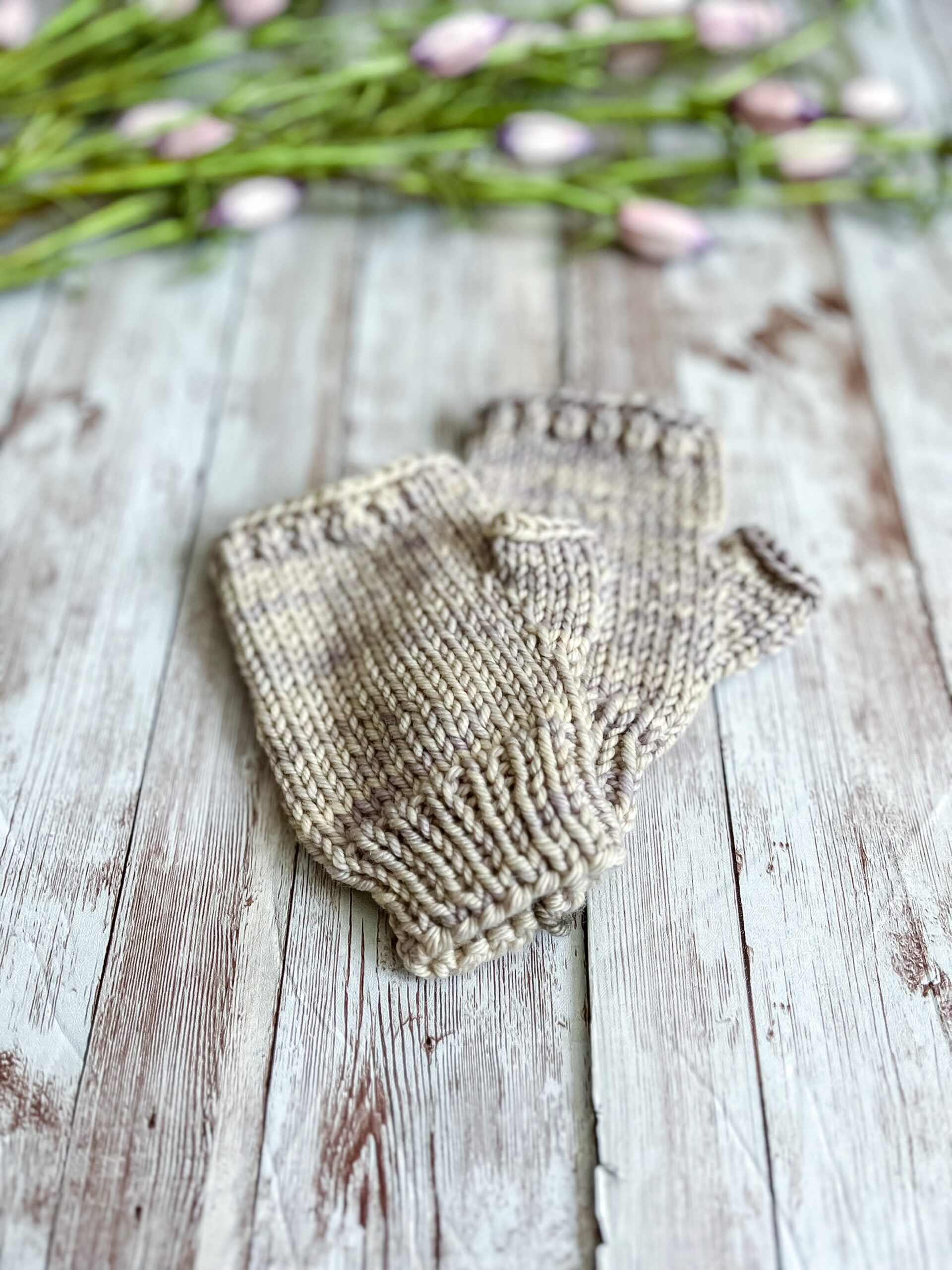 A pair of kid's sized light gray fingerless mitts are displayed on a wood plank with flower buds in the background