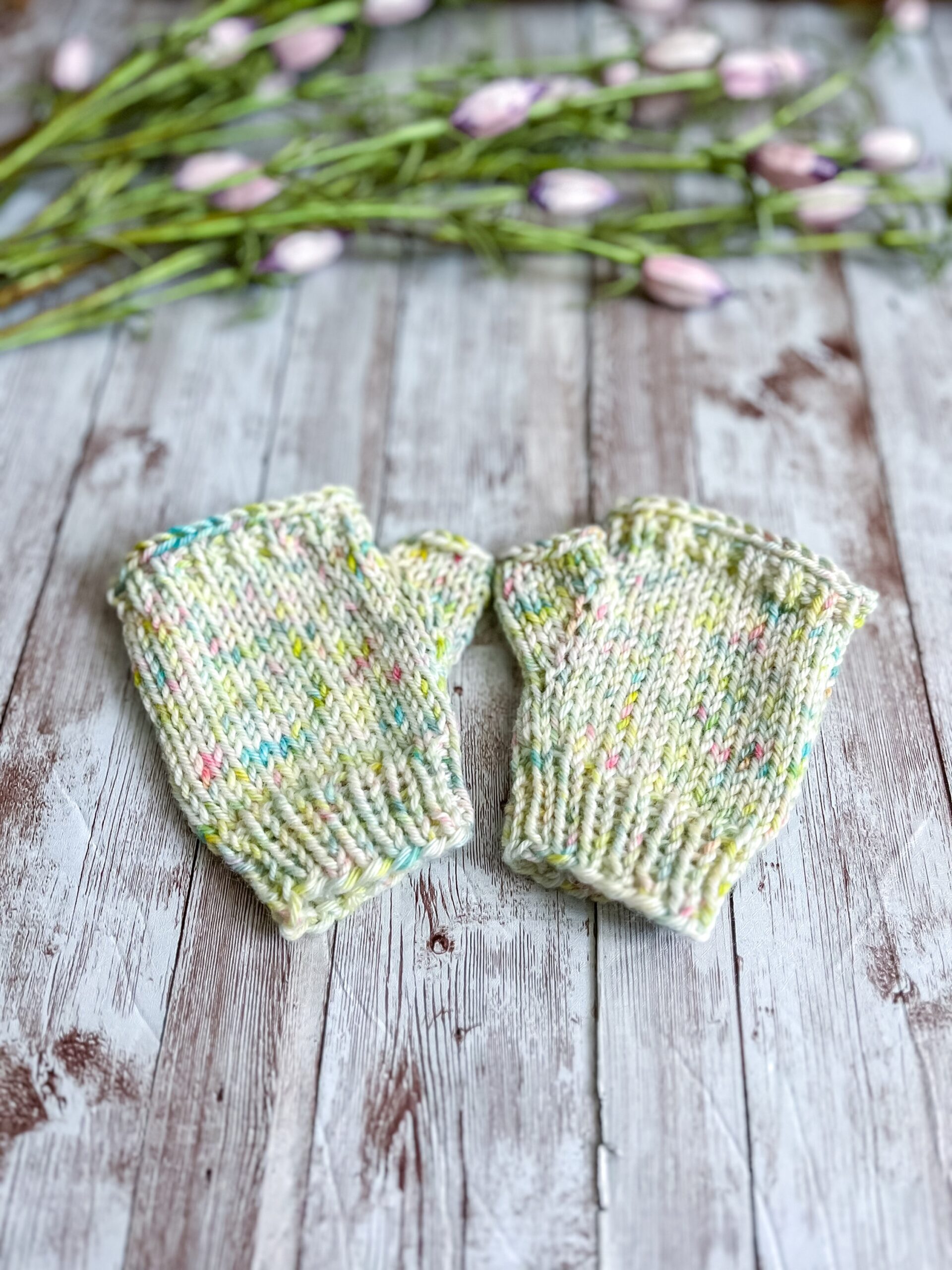 A pair of kid's sized pale yellow speckled fingerless mitts are displayed on a wood plank with flower buds in the background