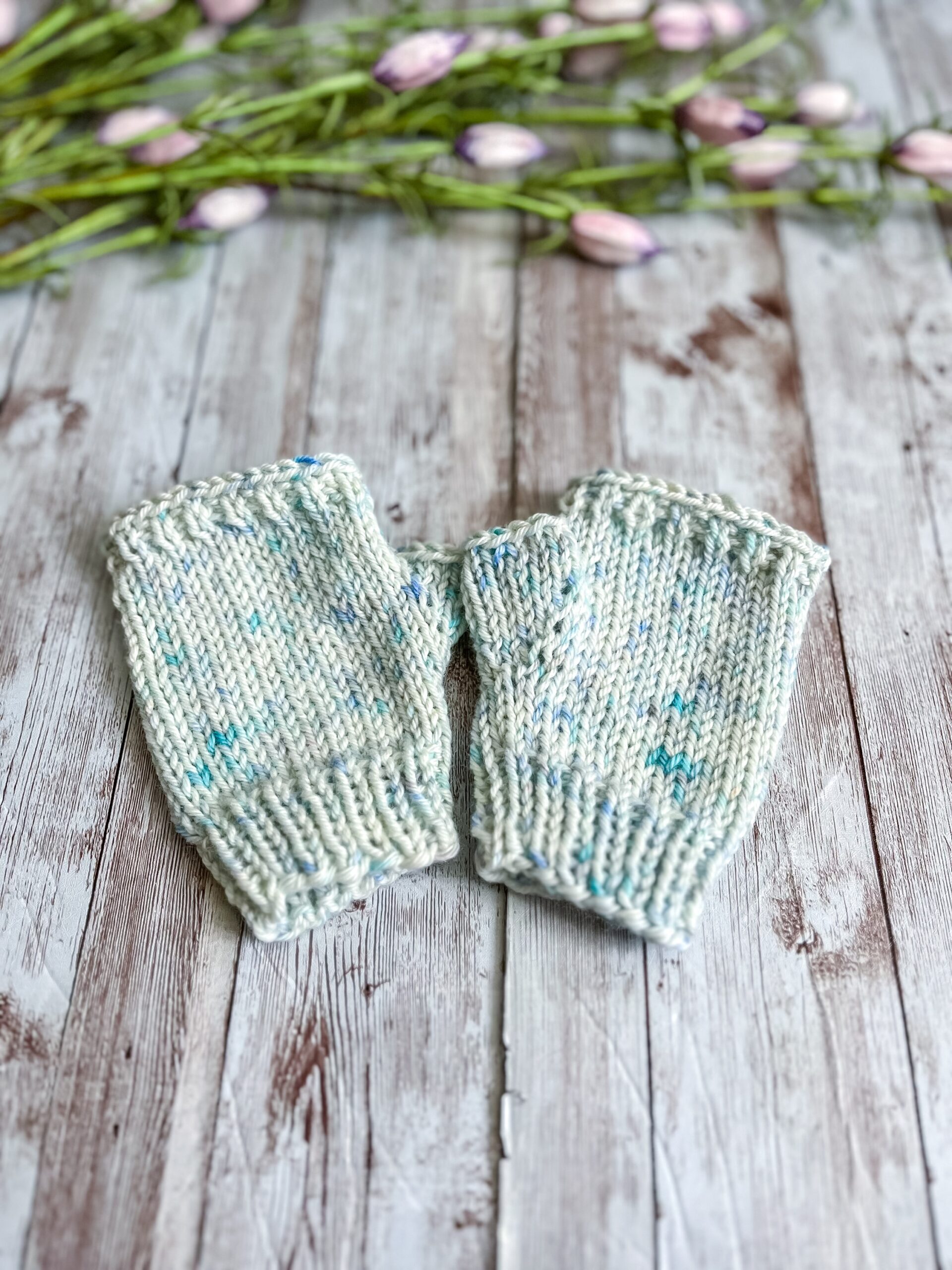 A pair of kid's sized pale blue speckled fingerless mitts are displayed on a wood plank with flower buds in the background