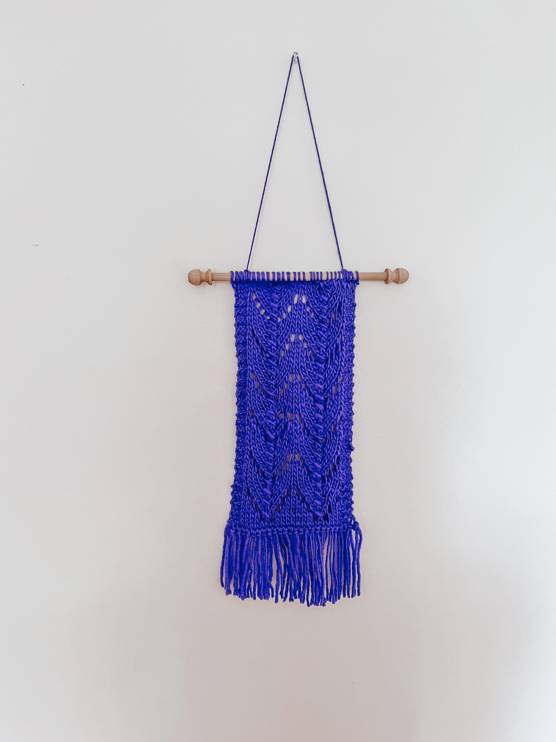 A periwinkle blue wool wall hanging is attached to a wooden dowel, hung from a longer string of wool. The hanging shows knitted chevrons in 2 columns with fringe on the bottom of the wall hanging.