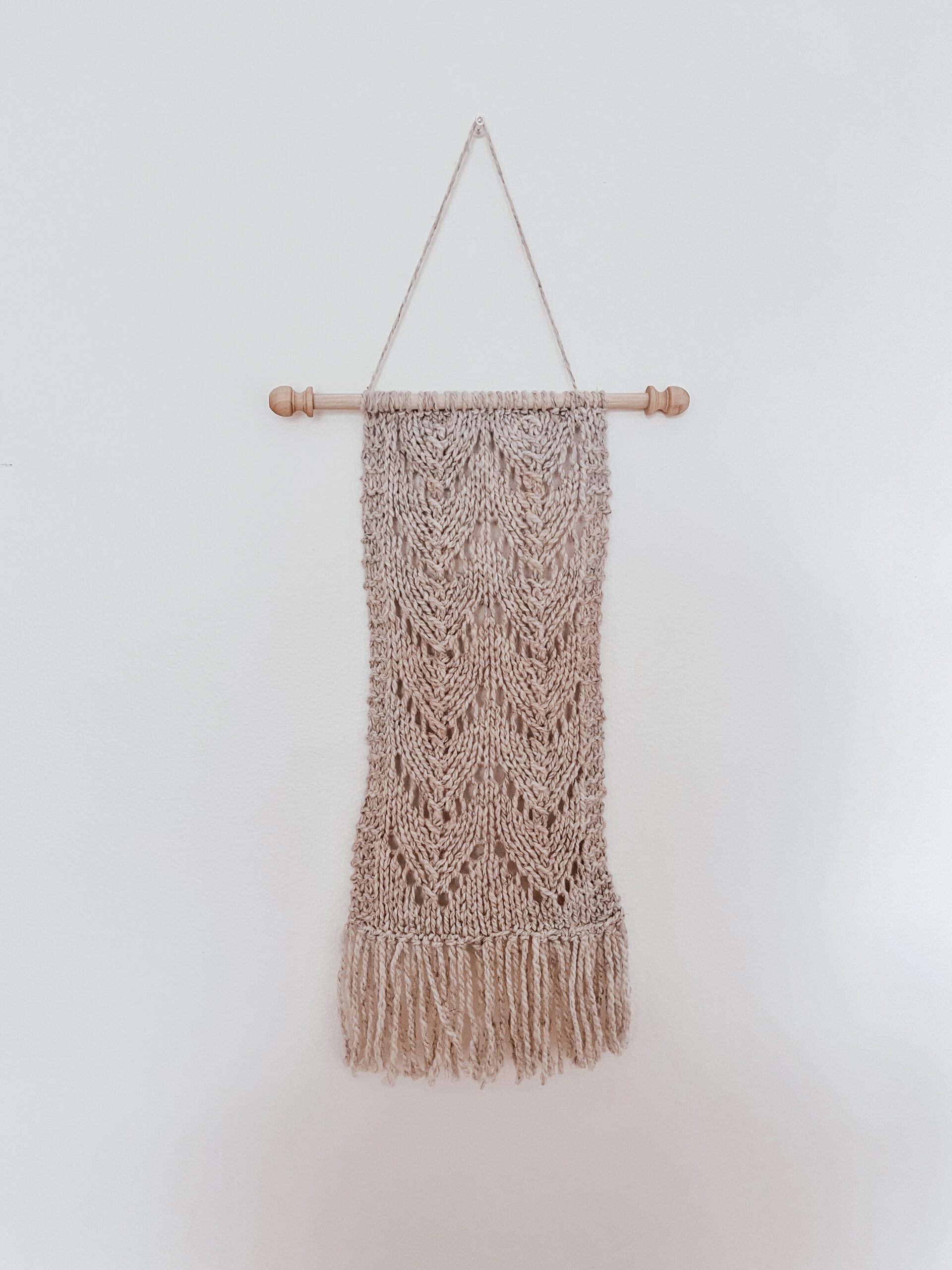 A natural tan wool wall hanging is attached to a wooden dowel, hung from a longer string of wool. The hanging shows knitted chevrons in 2 columns with fringe on the bottom of the wall hanging.