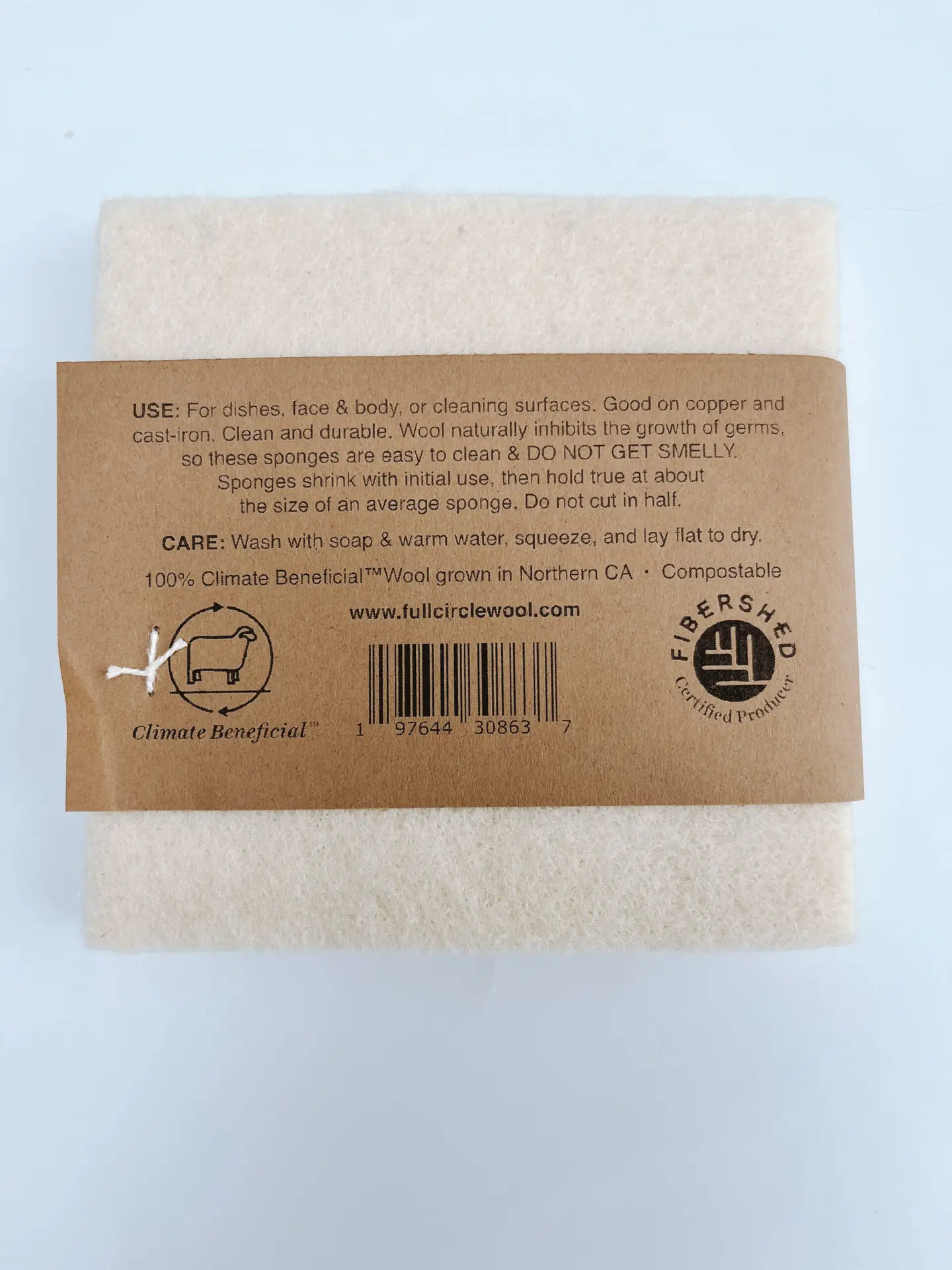 A set of 2 100% wool sponges is shown on a white background. The back label includes the text: "USE: for dishes, face & body, or cleaning surfaces. Good on copper and cast-iron. Clean and durable. Wool naturally inhibits the growth of germs so these sponges are easy to clean & DO NOT GET SMELLY. Sponges shrink with initial use, then hold true at about the size of an average sponge. Do not cut in half. CARE: Wash with soap & warm water, squeeze and lay flay to dry 100% Climate BeneficialTM Wool grown in Northern CA Compostable" It includes a logo of a sheep with the text Climate Beneficial and a logo of Fivershed Certified Producer