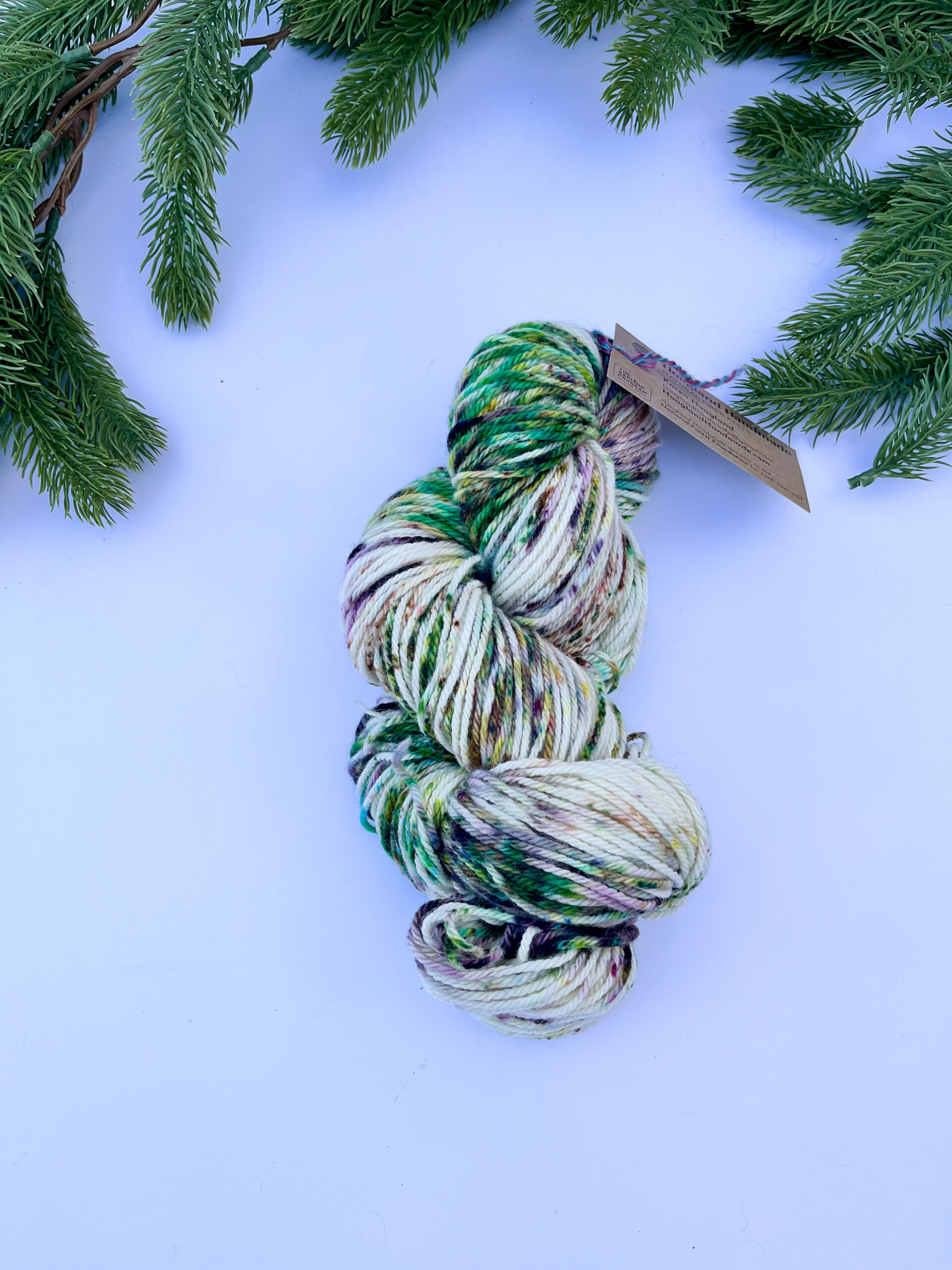 One skein of hand-dyed merino yarn is displayed on a white background with evergreen around the top border. The yarn features professional acid dye speckles in greens, burgundys, and browns.