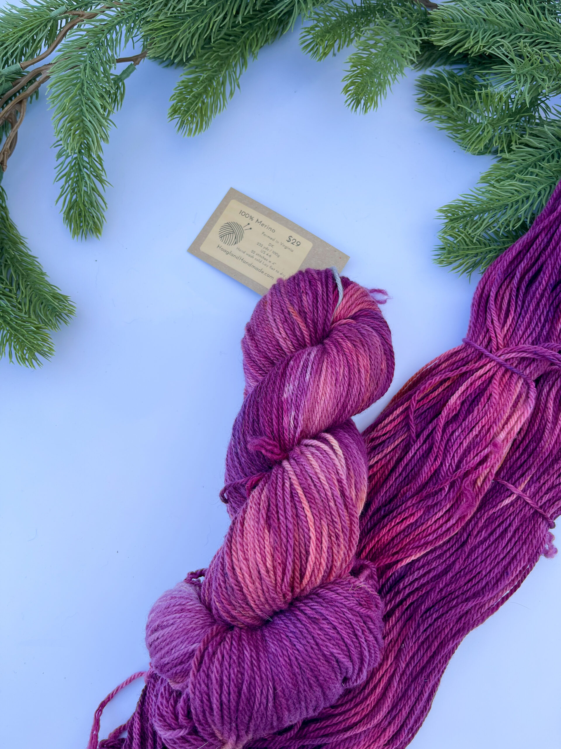 Two skeins of hand-dyed merino yarn are displayed on a white background with evergreen around the top border. The yarn features professional acid dyes in tonal cranberry, purple, and peach colors