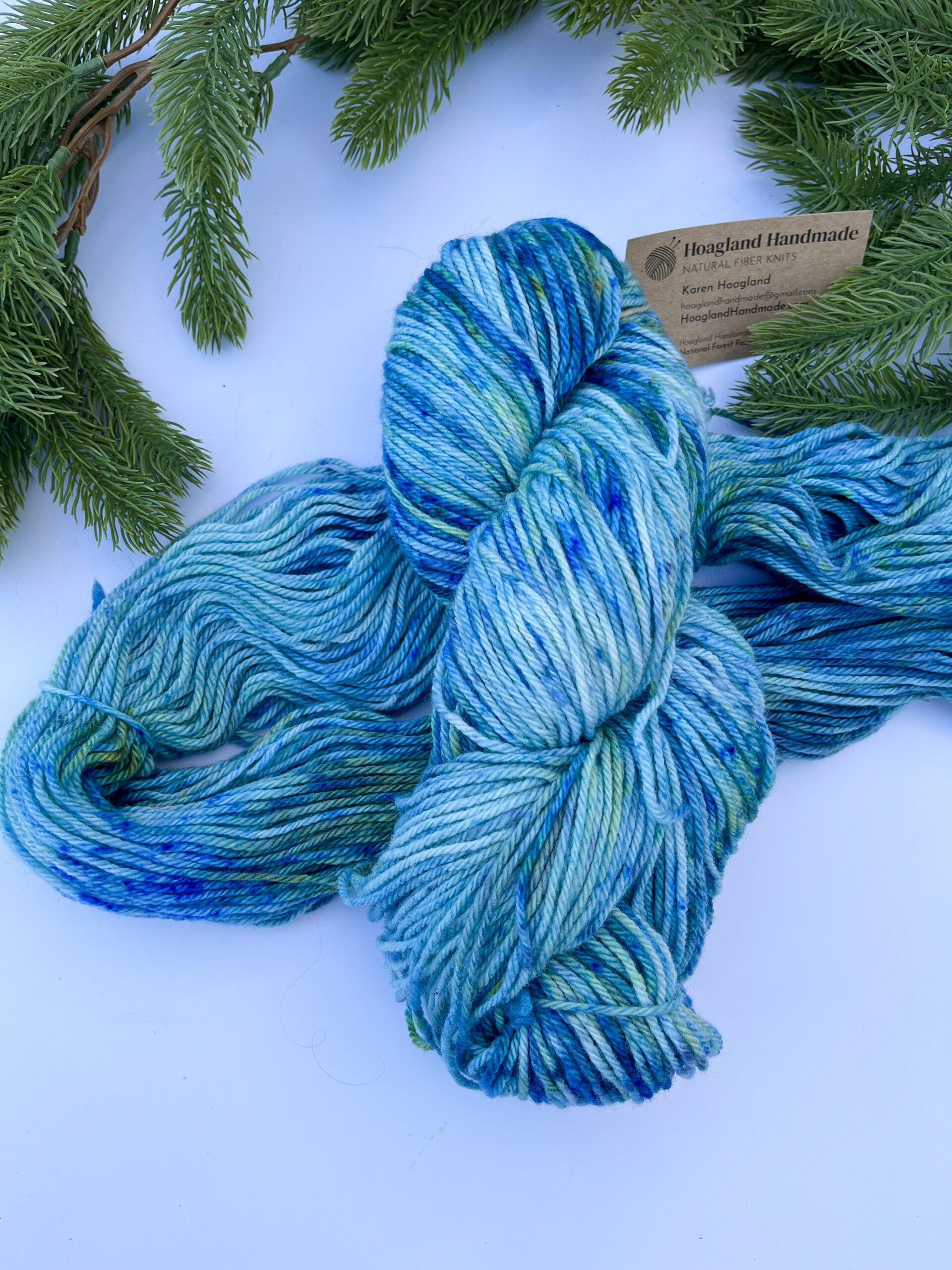 Two skeins of hand-dyed merino yarn are displayed on a white background with evergreen around the top border. The yarn features professional acid dyes in tonal blues with some green and gold speckles