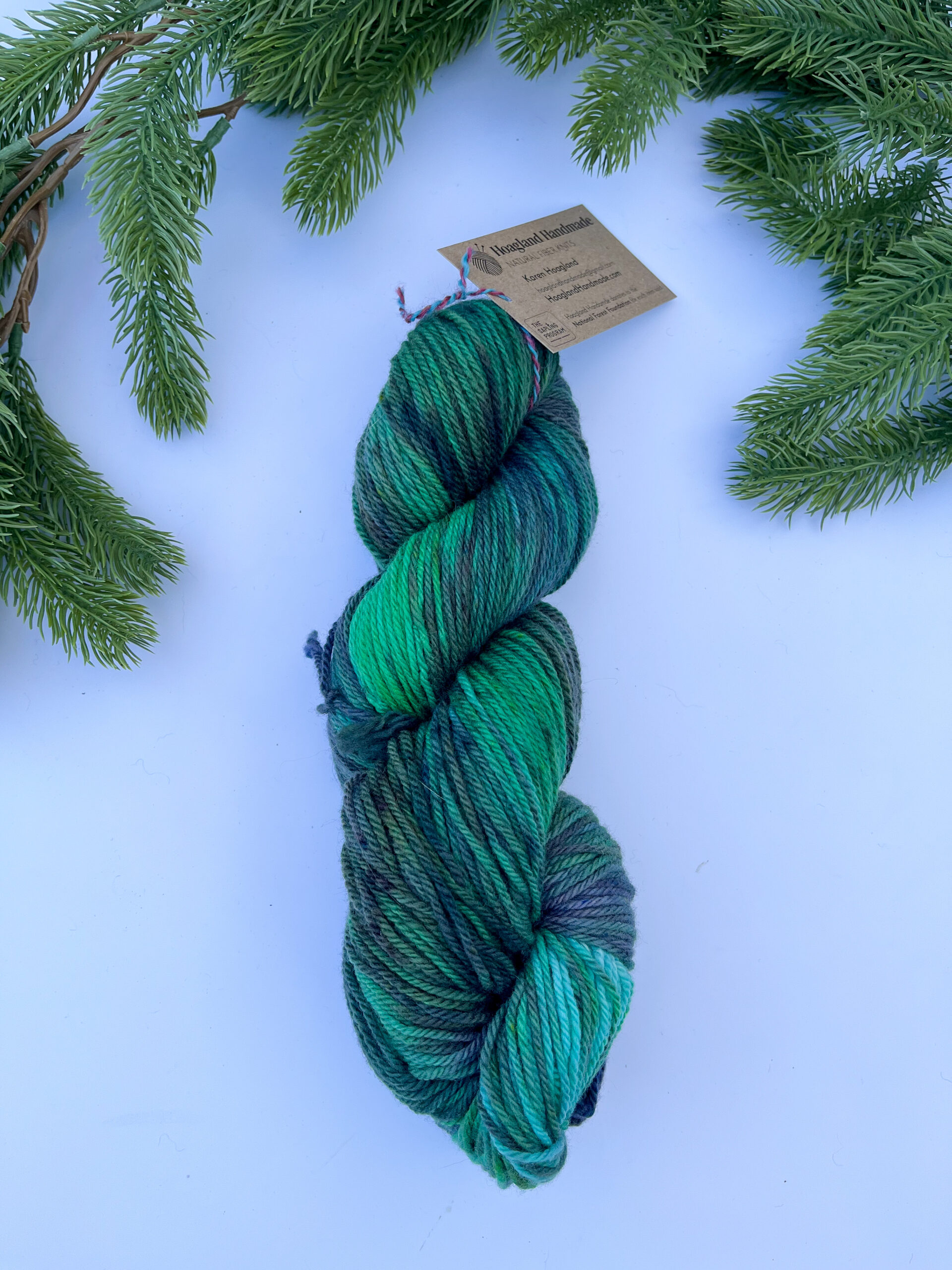 One skein of hand-dyed merino yarn is displayed on a white background with evergreen around the top border. The yarn features professional acid dyes in tonal greens and grays