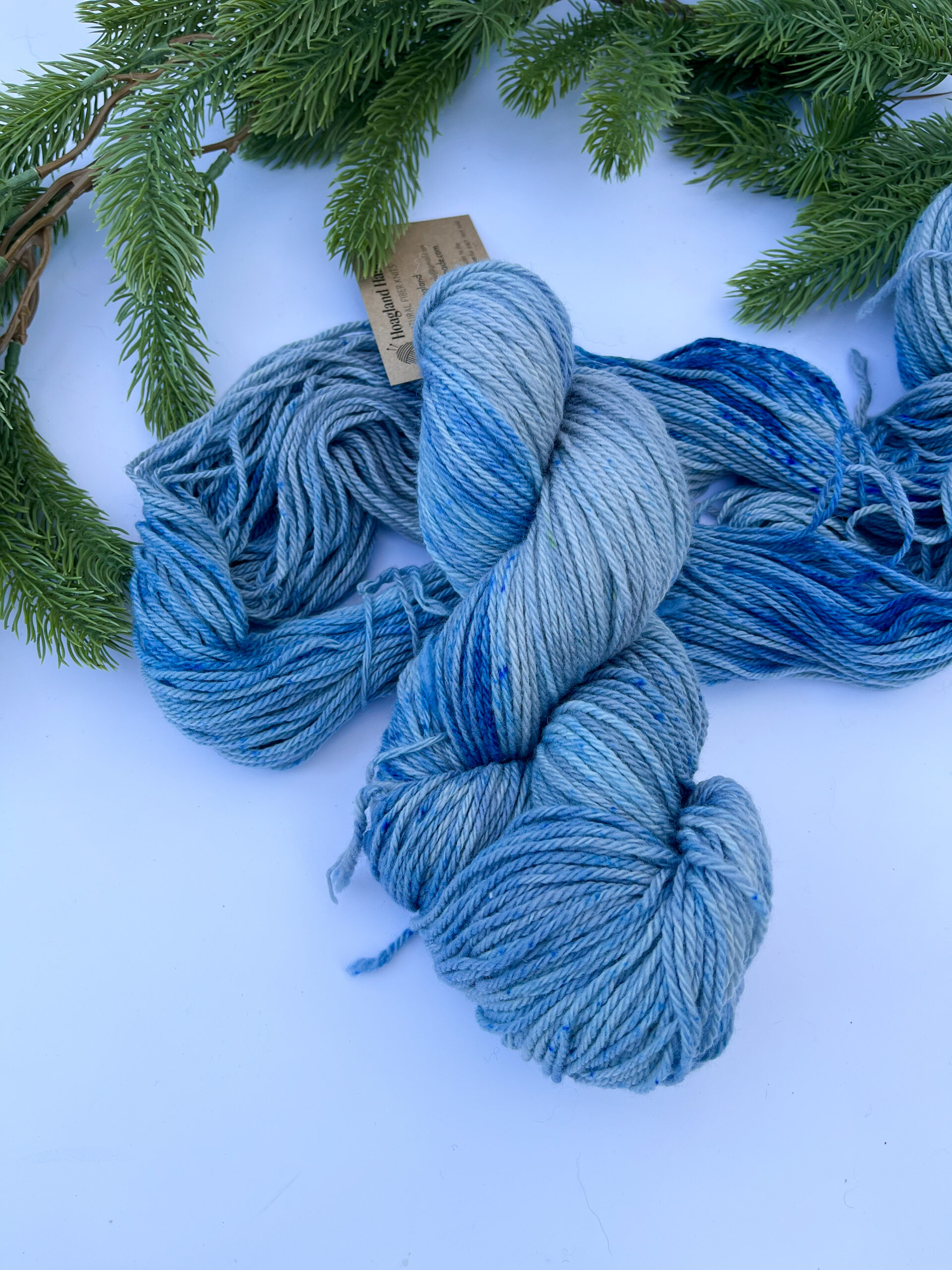 Two skeins of hand-dyed merino yarn are displayed on a white background with evergreen around the top border. The yarn features professional acid dyes in blues and grays