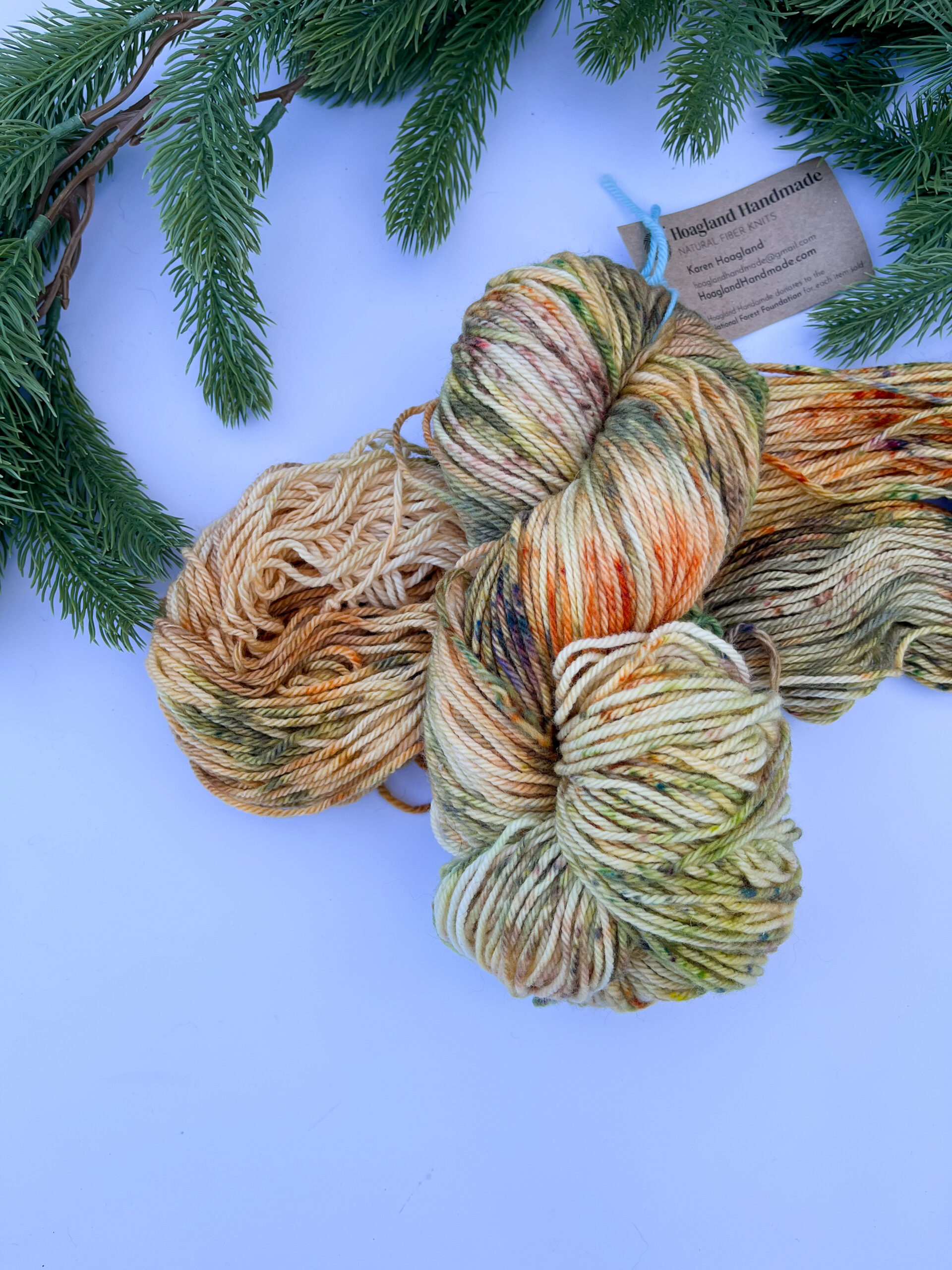 Two skeins of hand-dyed merino yarn are displayed on a white background with evergreen around the top border. The yarn features professional acid dyes in speckles oranges, greens, and browns